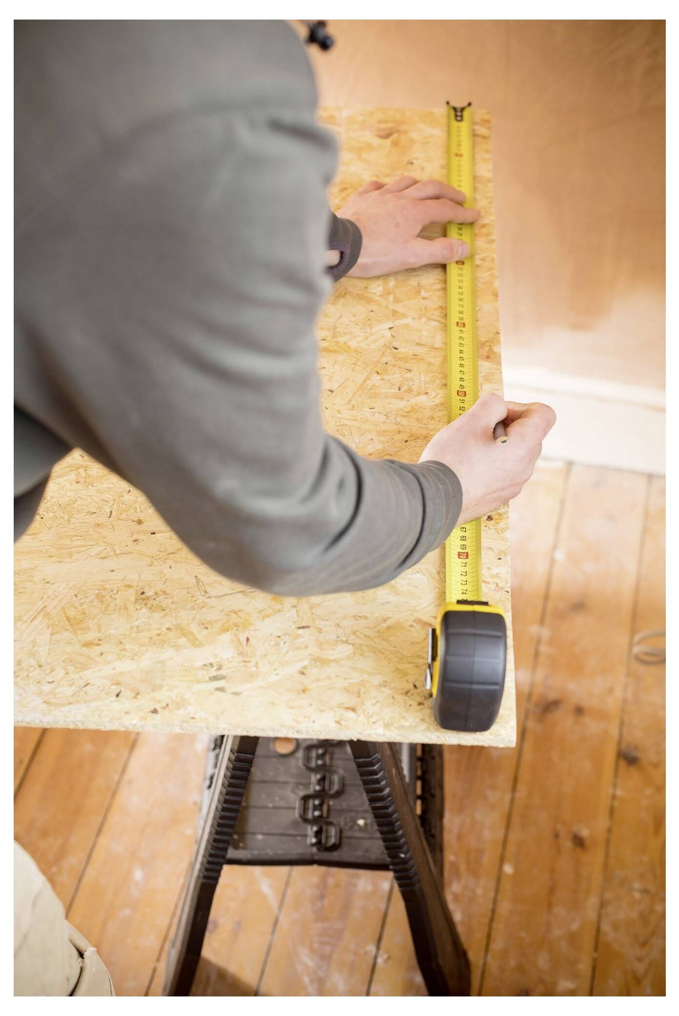 Eine Person misst ein Holzbrett mit einem gelben Stahllineal auf einem Sägebock und bereitet sich auf das Zuschneiden bei einer Schreinerei-Aufgabe vor.