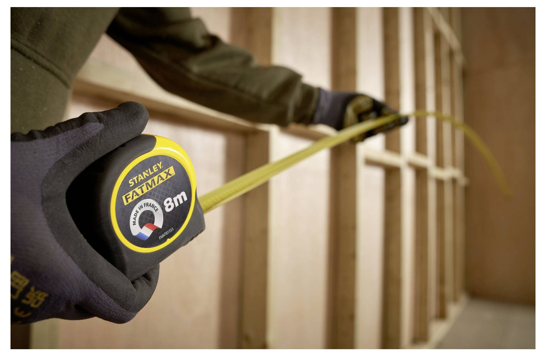 Eine Person mit Handschuhen hält ein ausgestrecktes 8-Meter-Stanley-Stahllineal gegen hölzerne Träger, was auf eine Bau- oder Mesaufgabe hindeutet.