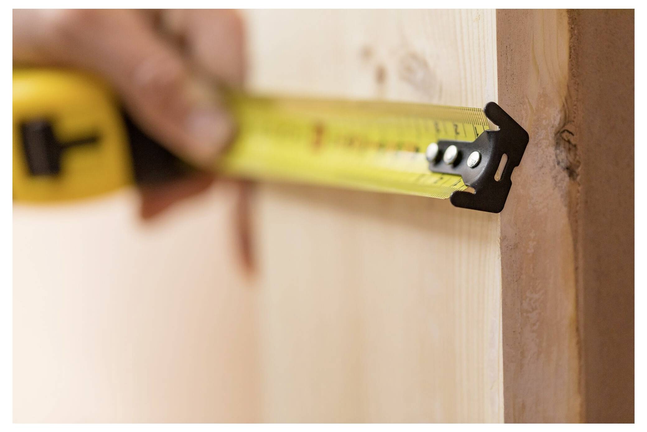 Eine Person hält ein gelbes Stahllineal in der Hand und misst die Dicke eines Holzbretts.