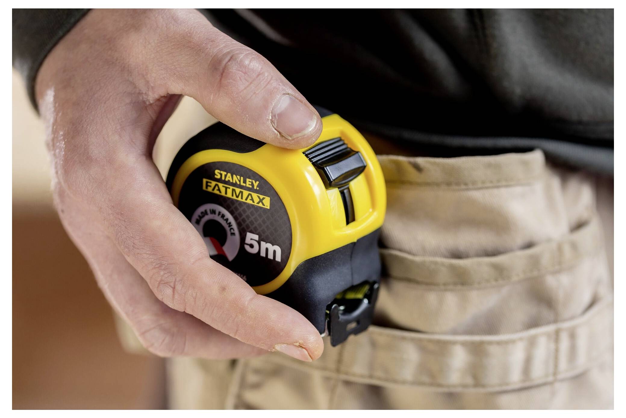 Eine Person befestigt ein gelbes 5-Meter-Stahllineal an einem beigen Werkzeuggürtel, um es einsatzbereit zu machen.