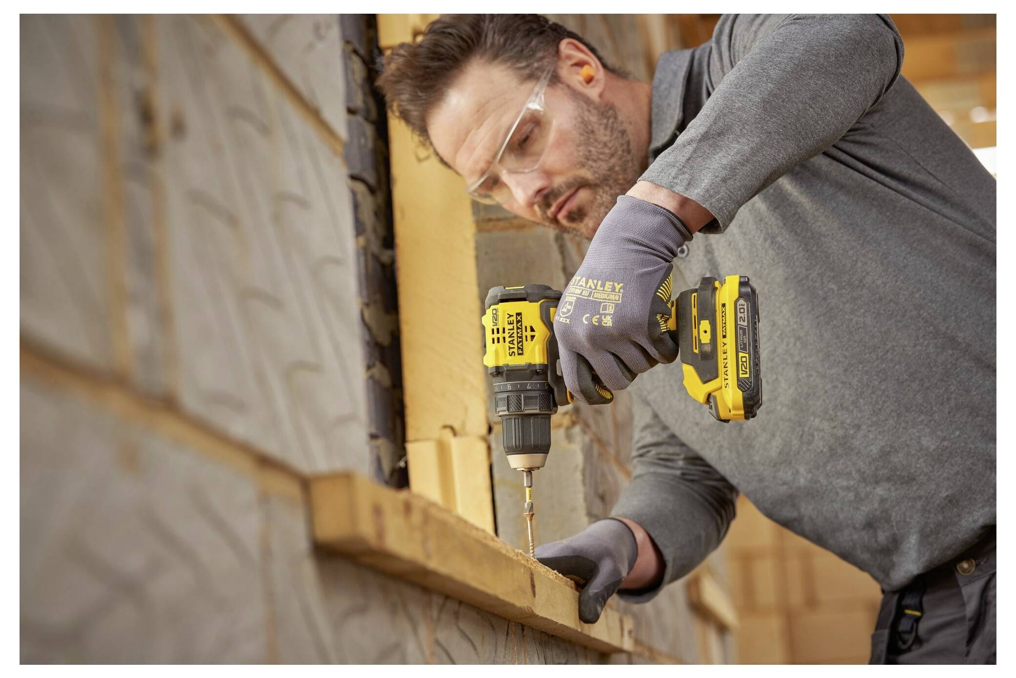 Ein Mann trägt eine Schutzbrille und Handschuhe und benutzt einen gelb-schwarz gestreiften Elektroschrauber, um eine Holzplatte auf einer Baustelle zu befestigen.