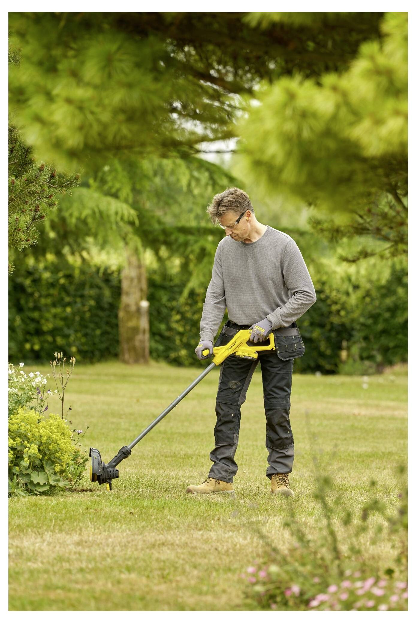 Eine Person trimmt das Gras mit einem gelben Fadentrimmer in einem Garten, der von Bäumen und Sträuchern umgeben ist.