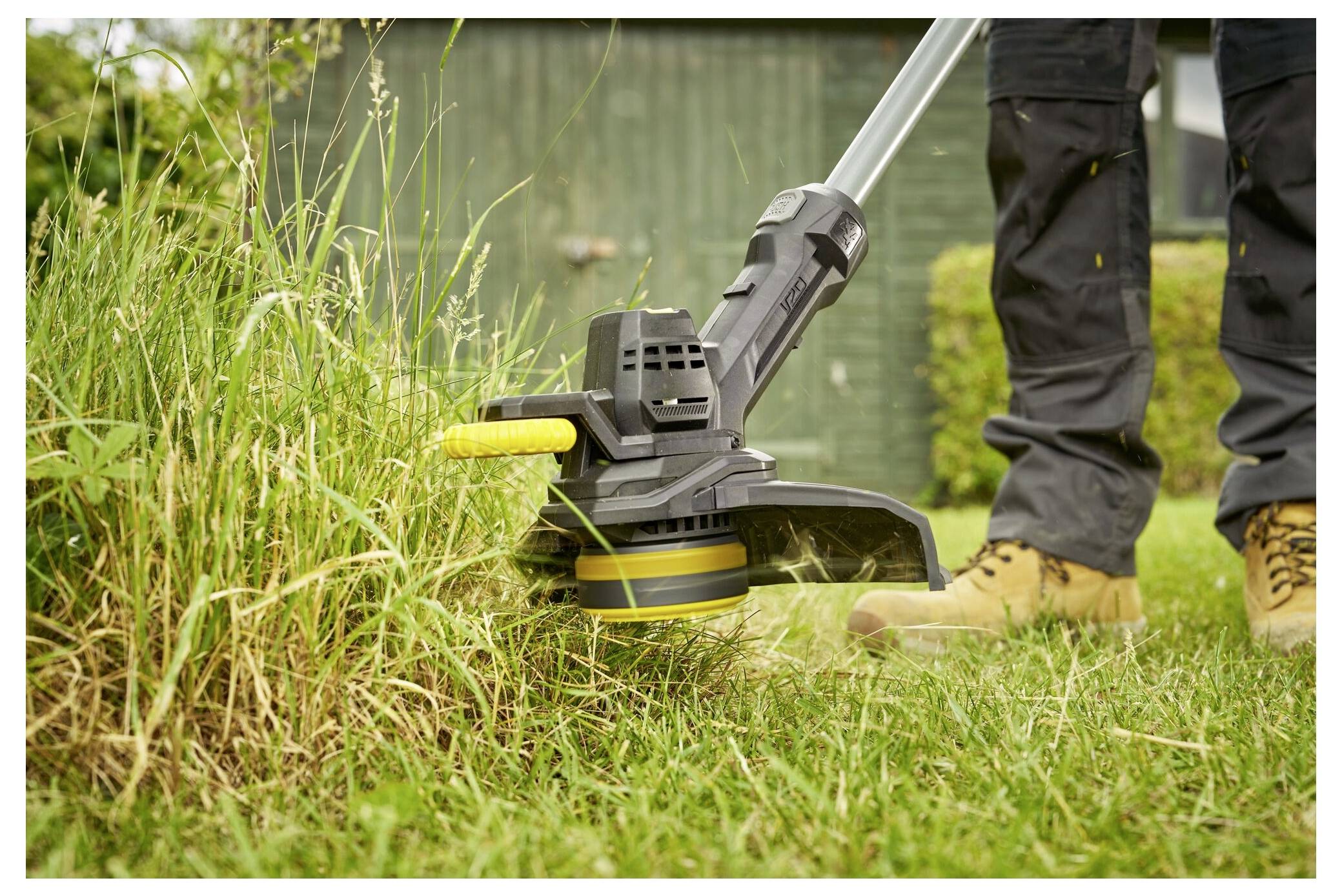 Eine Person mäht überwuchertes Gras mit einem gelb-schwarzen elektrischen Rasentrimmer in einem Garten und trägt dabei braune Stiefel und dunkle Hosen.
