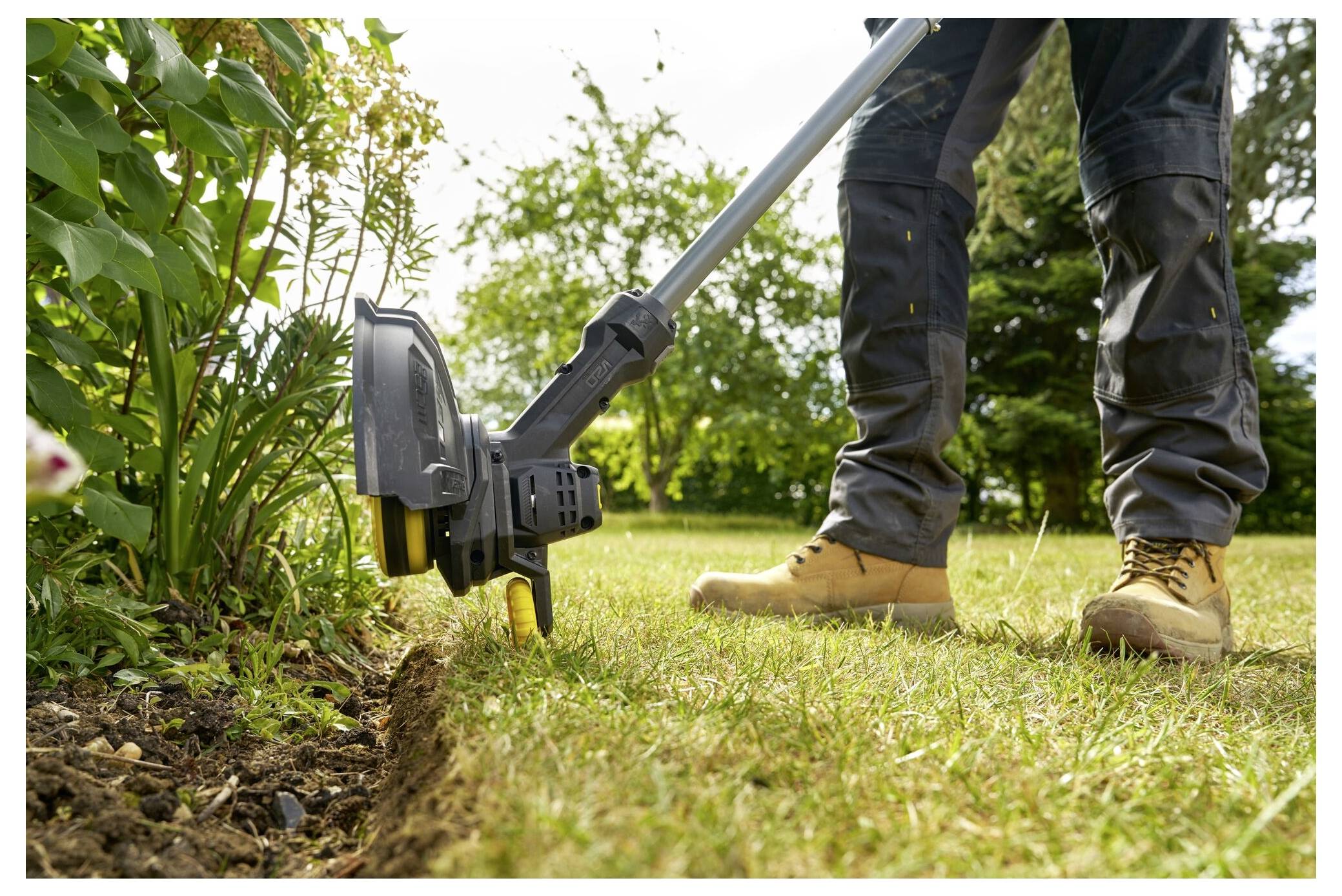Eine Person in Arbeitsstiefeln verwendet einen Kantenschneider, um die Kante eines Grasnestens neben einem Gartenbeet mit grünen Pflanzen zu trimmen.