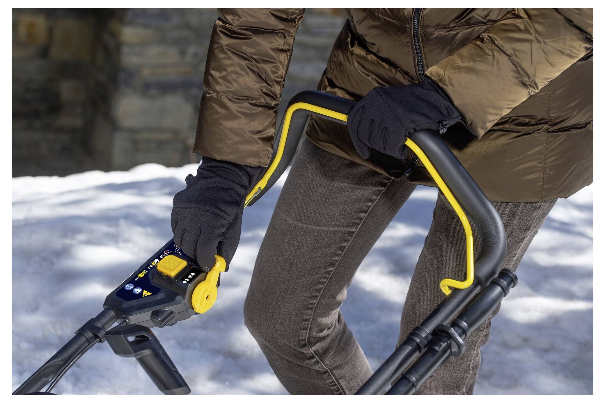 Eine Person, die eine braune Jacke und Handschuhe trägt, hält den Griff eines Schneefräse und steht auf Schnee.
