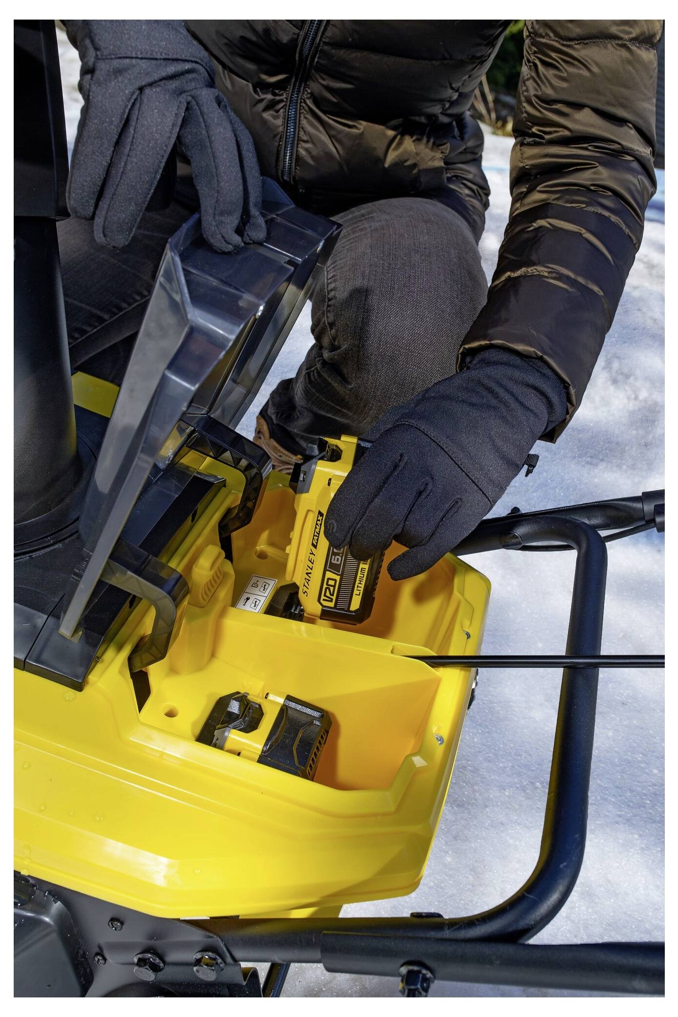 Eine Person in Winterkleidung setzt eine Batterie in einen gelben Schneefräser auf einer verschneiten Oberfläche ein.