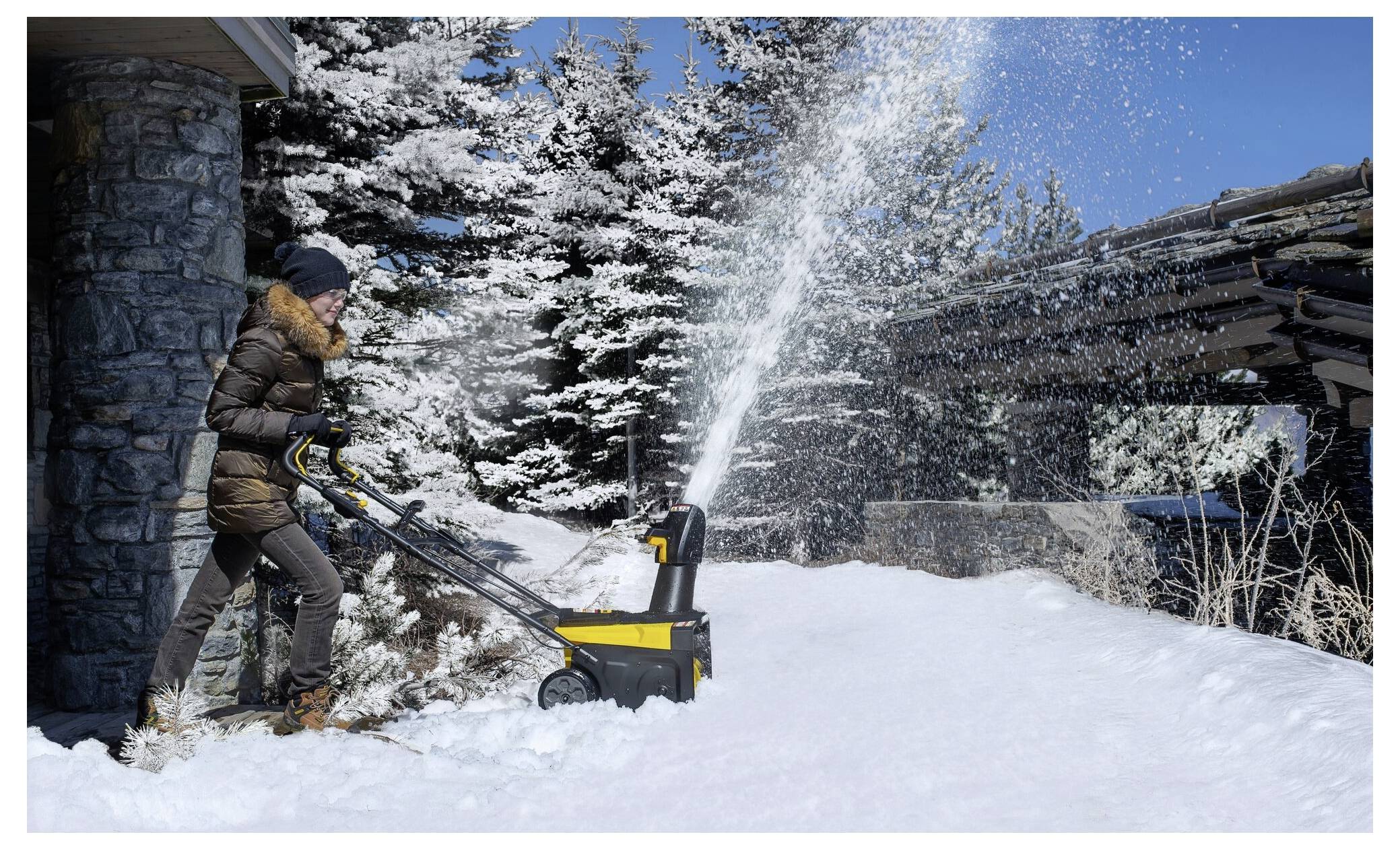 Eine Person in Winterkleidung verwendet einen Schneefräser, um einen verschneiten Weg zu räumen, der von Bäumen umgeben ist, mit einem klaren blauen Himmel im Hintergrund.