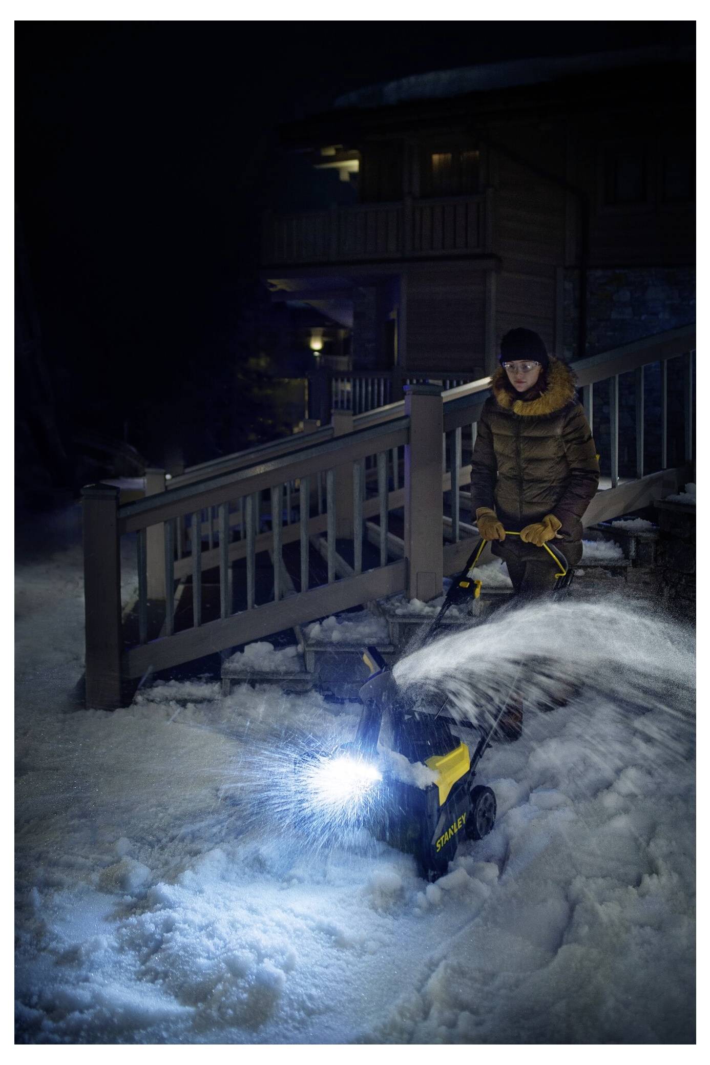 Eine Person bedient einen Schneefräser in der Nacht und räumt Schnee von einem Weg vor einem beleuchteten Holzhaus.