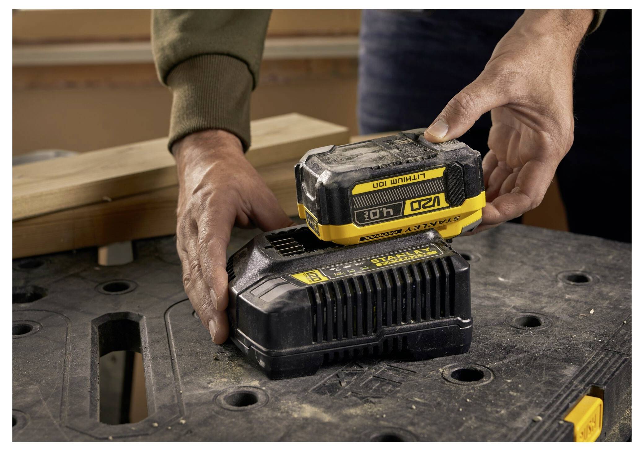 Hände legen ein wiederaufladbares Batteriepaket auf eine Ladestation auf einer Werkbank, mit Holzbrettern in der Nähe.