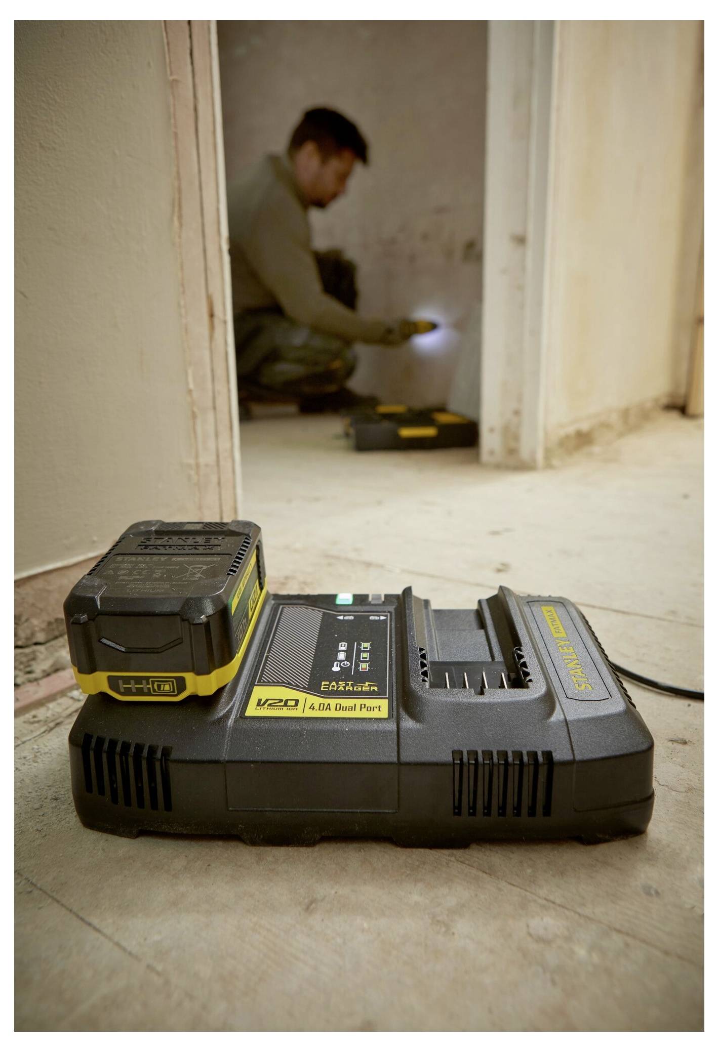 Ein schwarzes und gelbes Ladegerät sitzt auf einem Boden; eine Person im Hintergrund kniet und benutzt eine Taschenlampe in einem Raum.
