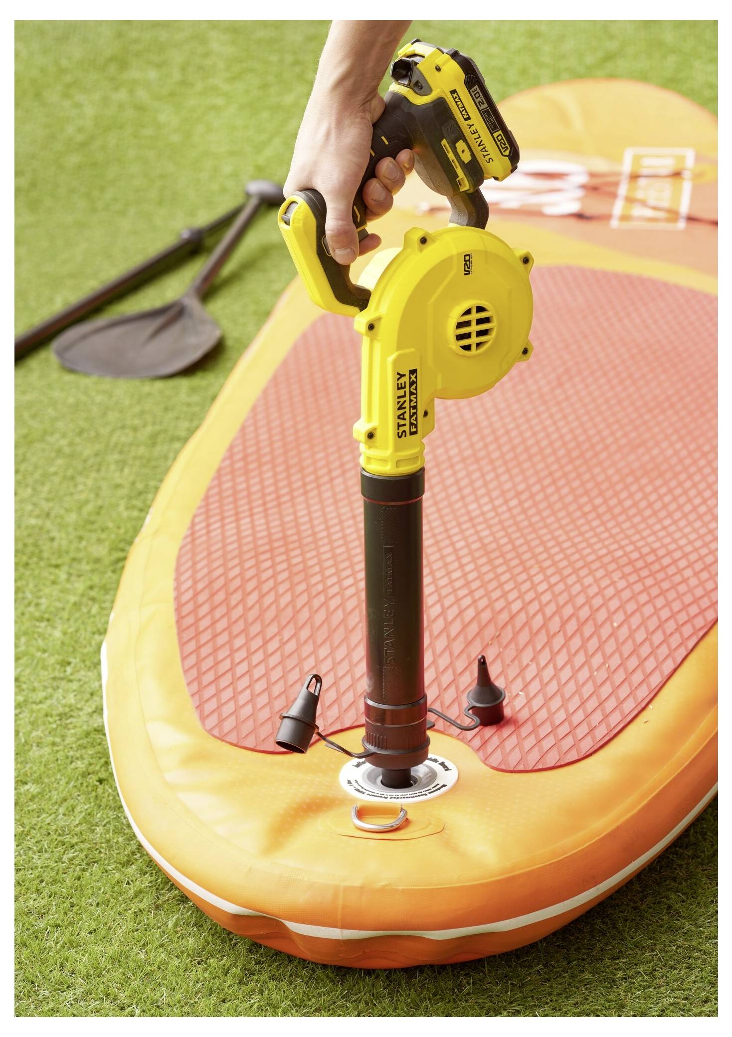 Eine Person bläst ein orangefarbenes Paddleboard mit einer gelben elektrischen Pumpe auf, während ein Paddel in der Nähe auf dem Gras liegt.