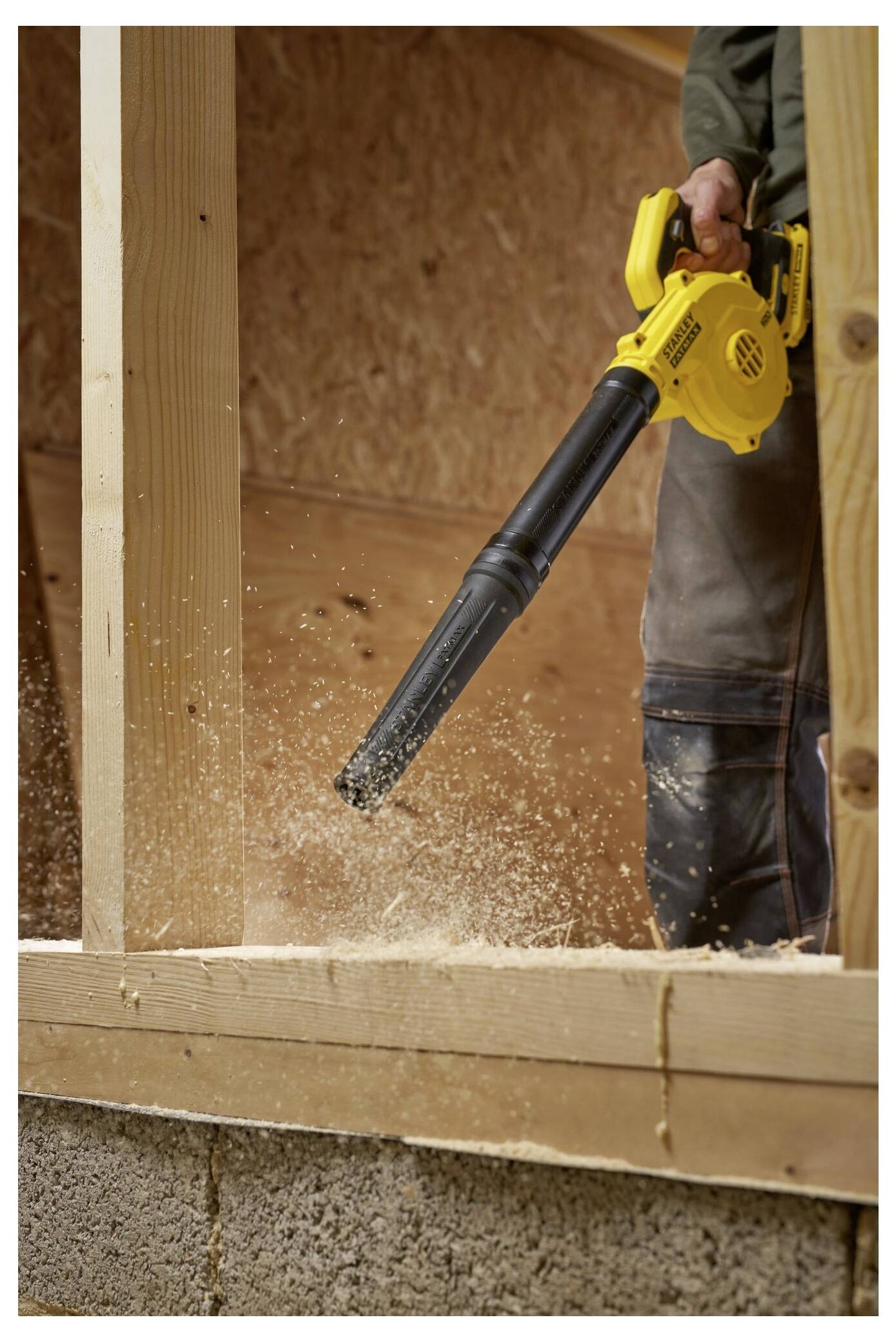 Eine Person benutzt einen gelben Laubbläser, um Sägespäne von Holzbrettern zu entfernen, wobei Holzspäne in einer unfertigen Struktur sichtbar fliegen.