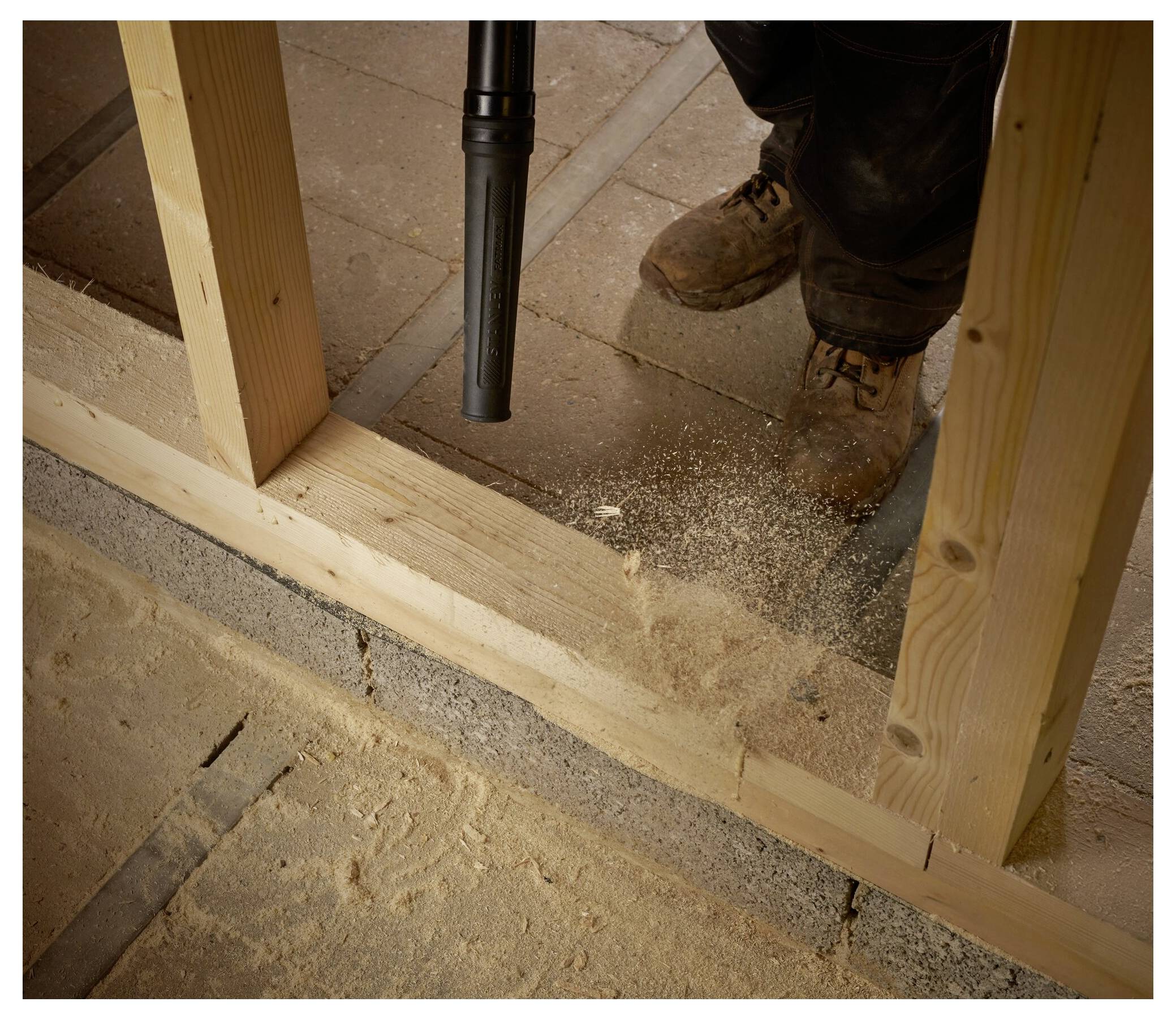 Ein Arbeiter saugt Sägespäne von einem Baustellenboden auf, umgeben von Holzrahmen und laufenden Baumaterialien.