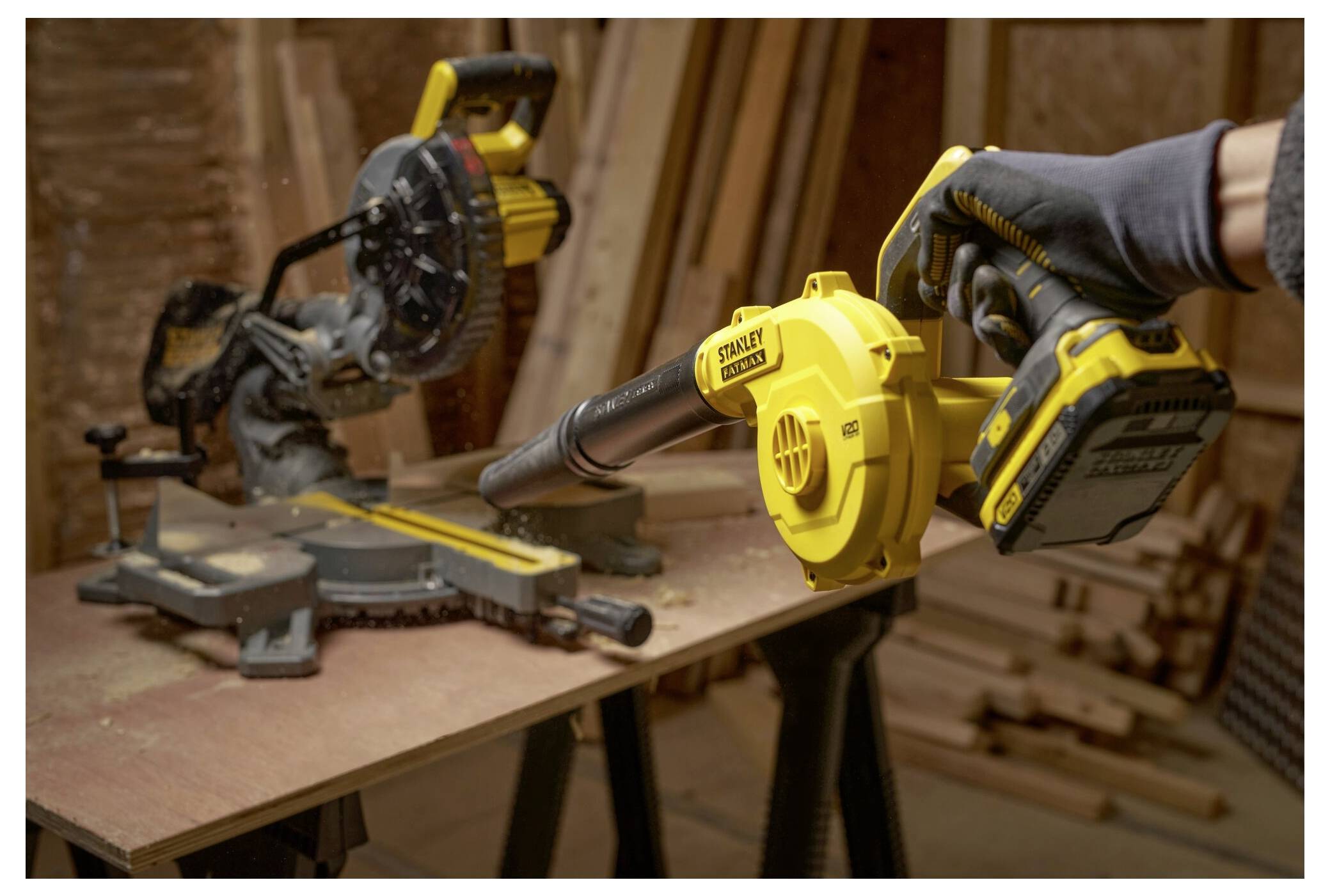 Eine Person nutzt ein tragbares Gebläse, um Sägespäne von einer Werkbank in einer Holzwerkstatt zu entfernen. Eine Gehrungssäge und Holzbretter befinden sich im Hintergrund.