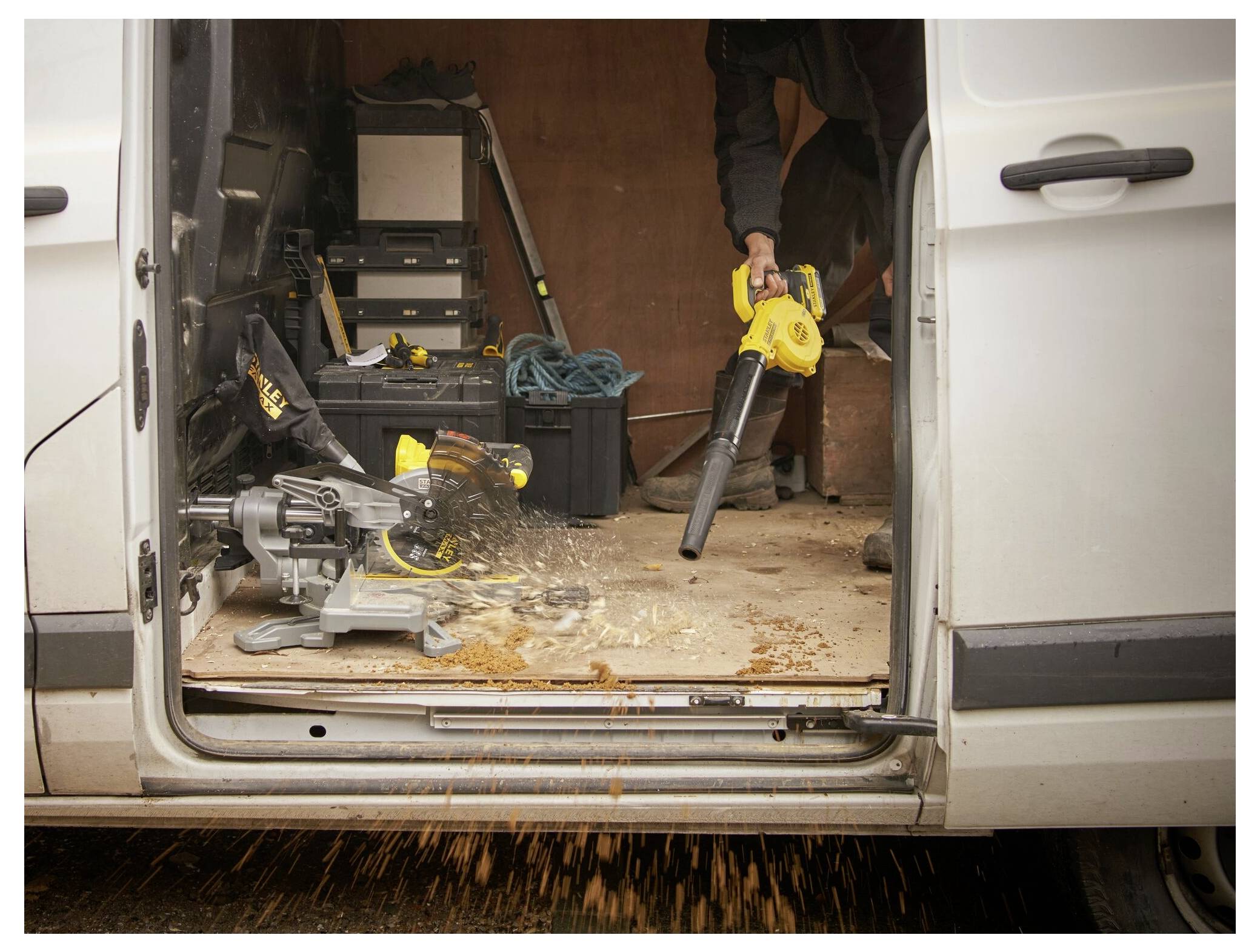 Zwei behandschuhte Hände halten ein gelb-schwarzes Stanley FatMax Multitool, das verschiedene Aufsätze und ein Stromkabel auf einer Holzoberfläche zeigt.
