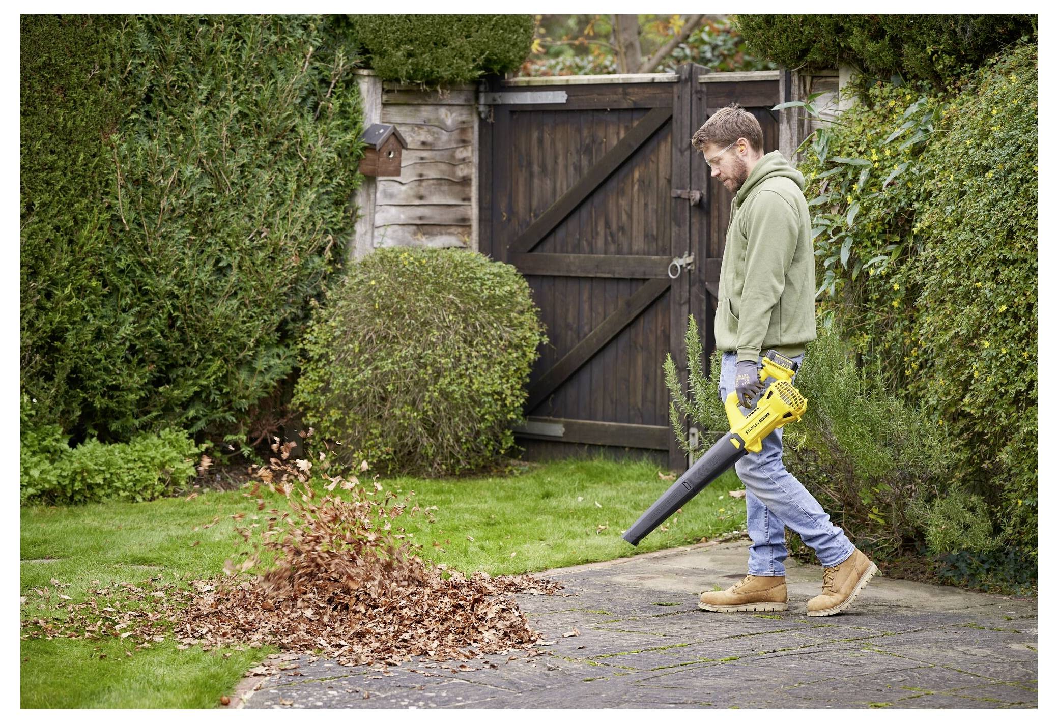 Eine Person, die einen Laubbläser benutzt, um gefallene Blätter von einem Gartenweg zu entfernen, steht in der Nähe eines geschlossenen Holztors, das von Grünanlagen umgeben ist.