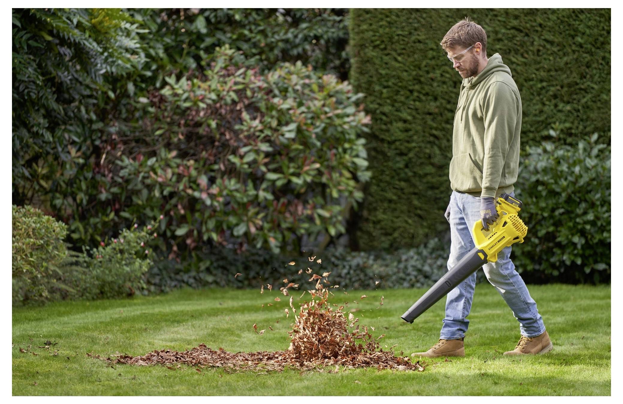 Ein Mann benutzt eine gelbe Stanley-Bohrmaschine, um Metallplatten in Holzbalken in einem unfertigen Haus einzuschrauben. Er trägt eine Schutzbrille und Handschuhe.