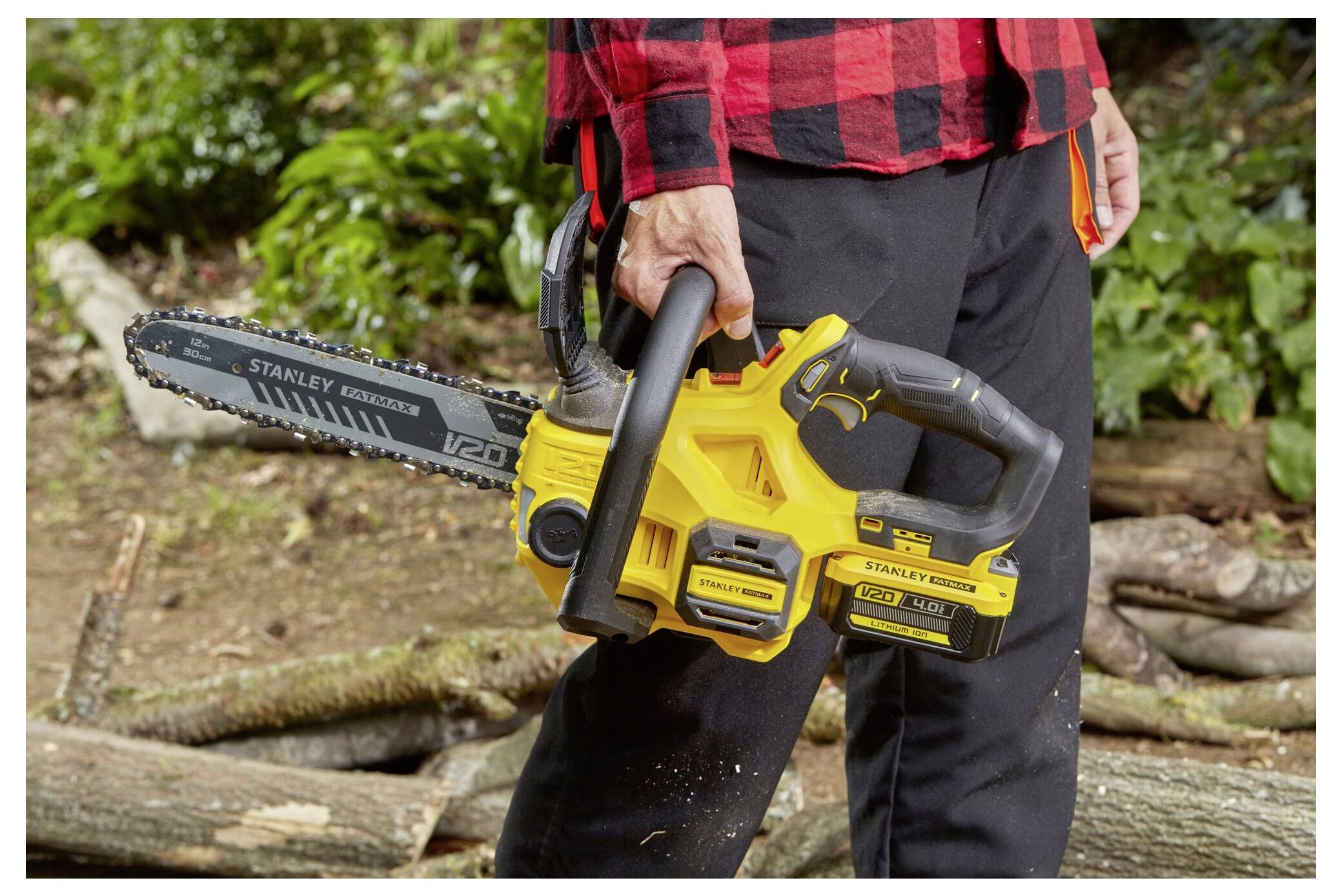 Eine Person in einem roten karierten Hemd und schwarzen Hosen hält eine gelbe Kettensäge in einem bewaldeten Gebiet mit Baumstämmen und Blättern auf dem Boden.