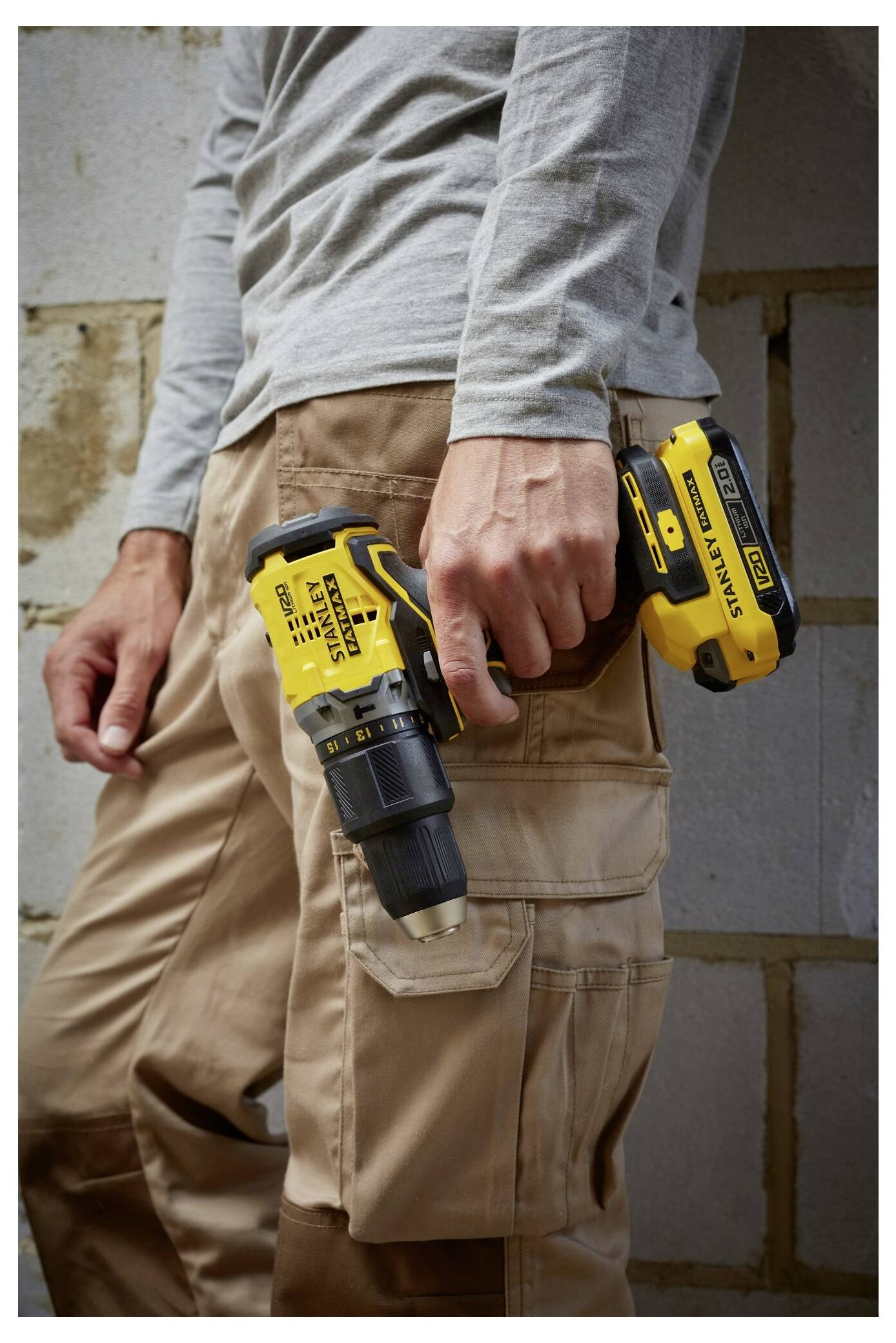 Eine Person, die khakifarbene Hosen und ein graues Langarmhemd trägt, hält einen gelb-schwarzen kabellosen Bohrer in der Hand und steht vor einer Backsteinmauer.