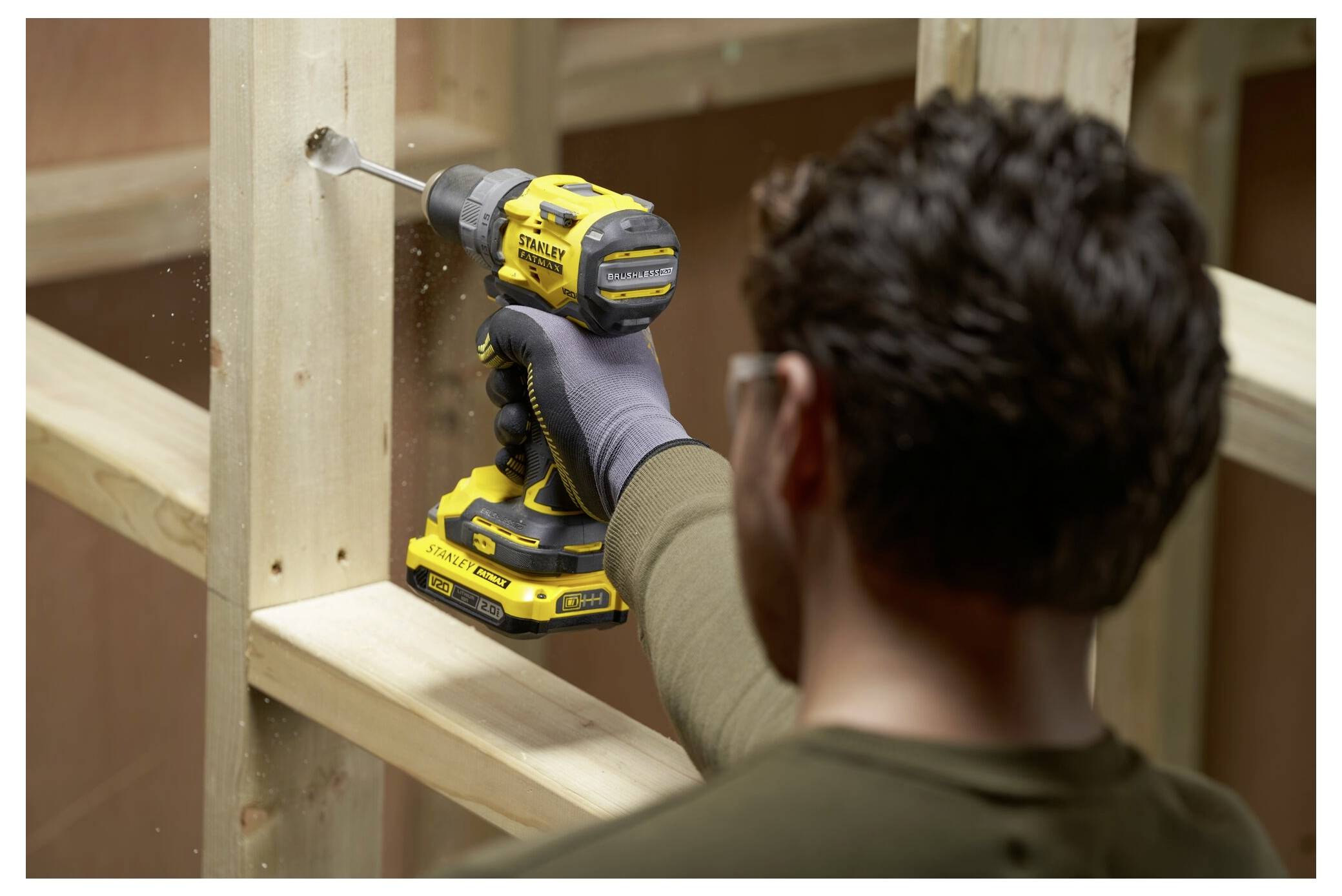 Eine Person benutzt einen gelben kabellosen Bohrer, um ein Loch in einen Holzbalken innerhalb eines Baurahmens zu bohren, wobei sie eine Schutzbrille und Handschuhe trägt.