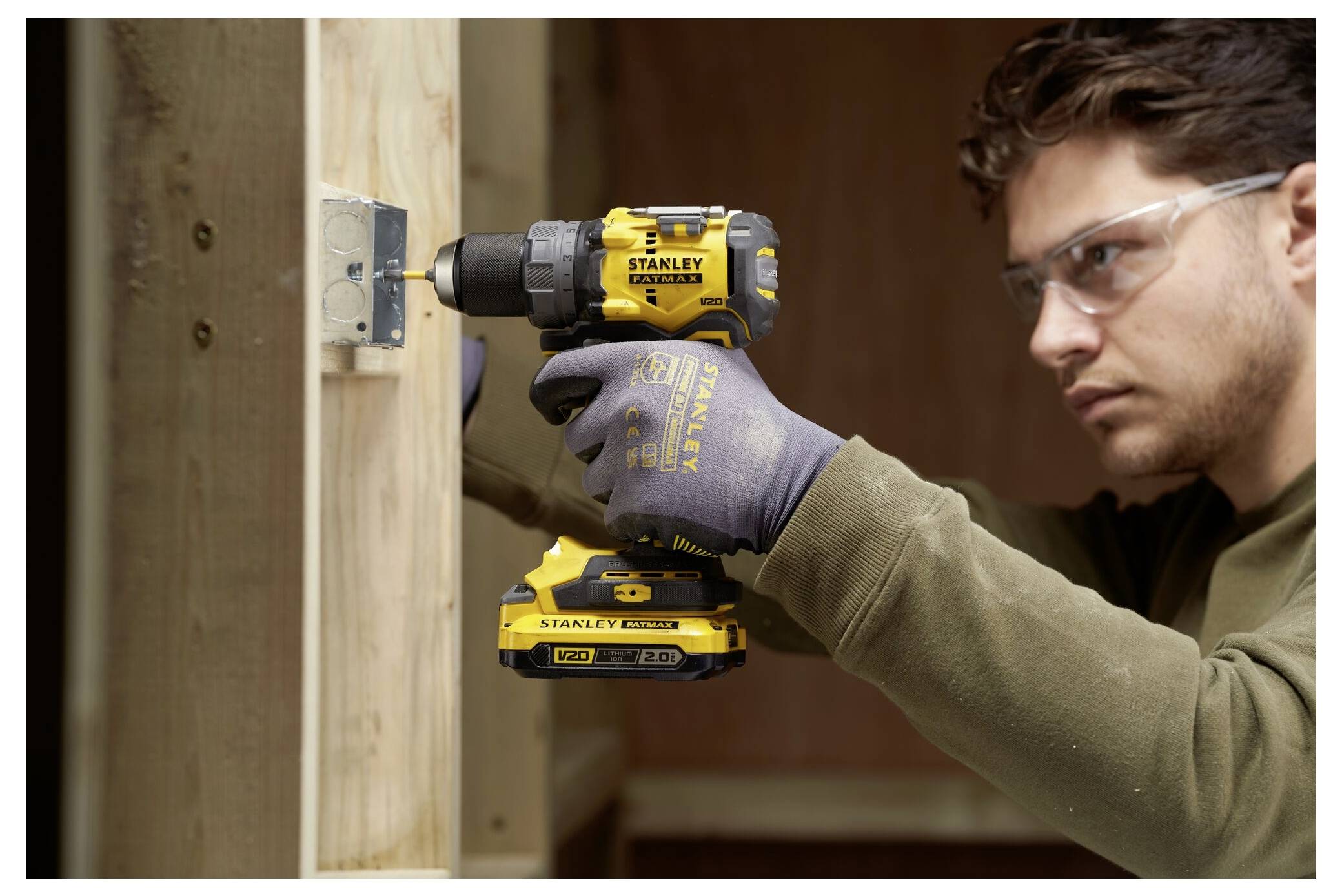 Eine Person verwendet einen gelb-schwarzen Elektroschrauber, um eine Metallhalterung an einer Holzstruktur zu befestigen, während sie eine Schutzbrille und Handschuhe trägt.