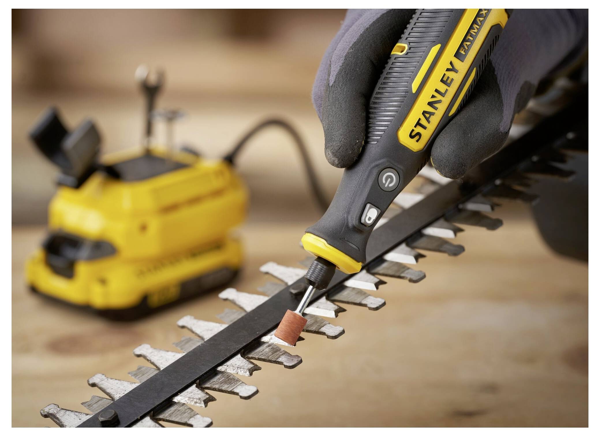 Eine Hand benutzt ein Stanley-Elektroschleifsystem, um die Klinge einer Heckenschere zu schärfen. Ein gelbes Elektrowerkzeug ist im Hintergrund sichtbar.