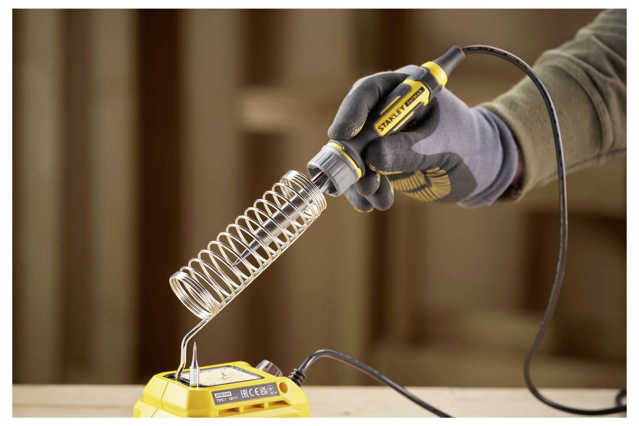 Eine behandschuhte Hand hält einen Lötkolben über seinen Ständer auf einer Werkbank, mit einem Holzhintergrund.