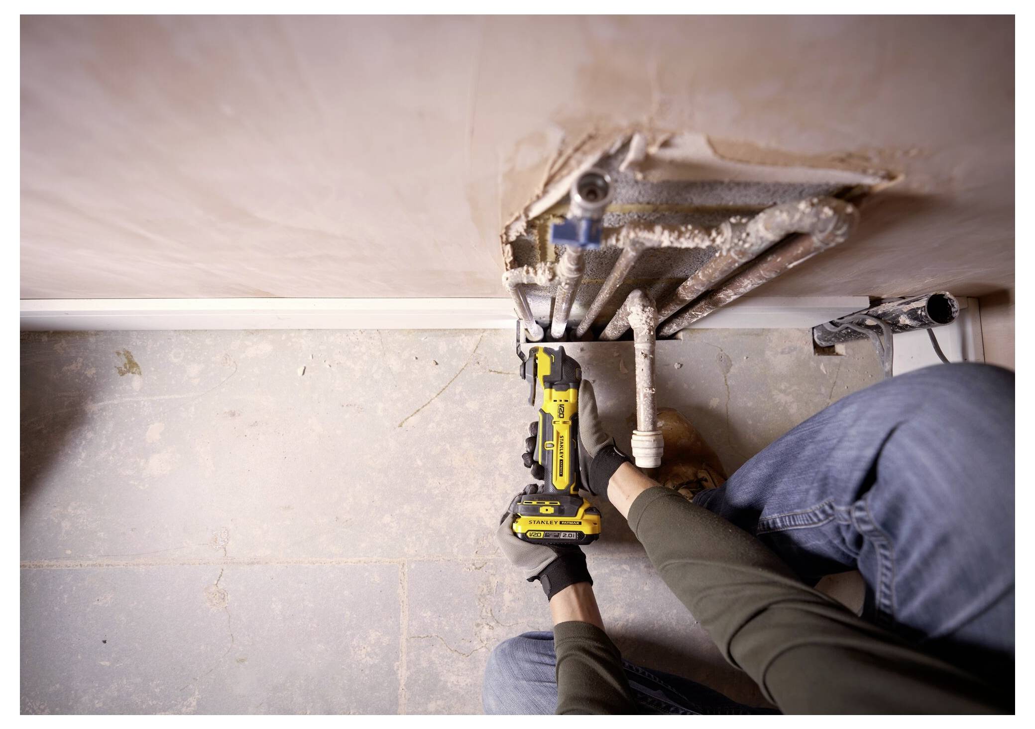 Eine Person in Handschuhen verwendet ein gelbes schnurloses Werkzeug zur Reparatur von freiliegenden Rohren neben einer Wand. Der Boden ist unfertig mit Staub und Schutt.