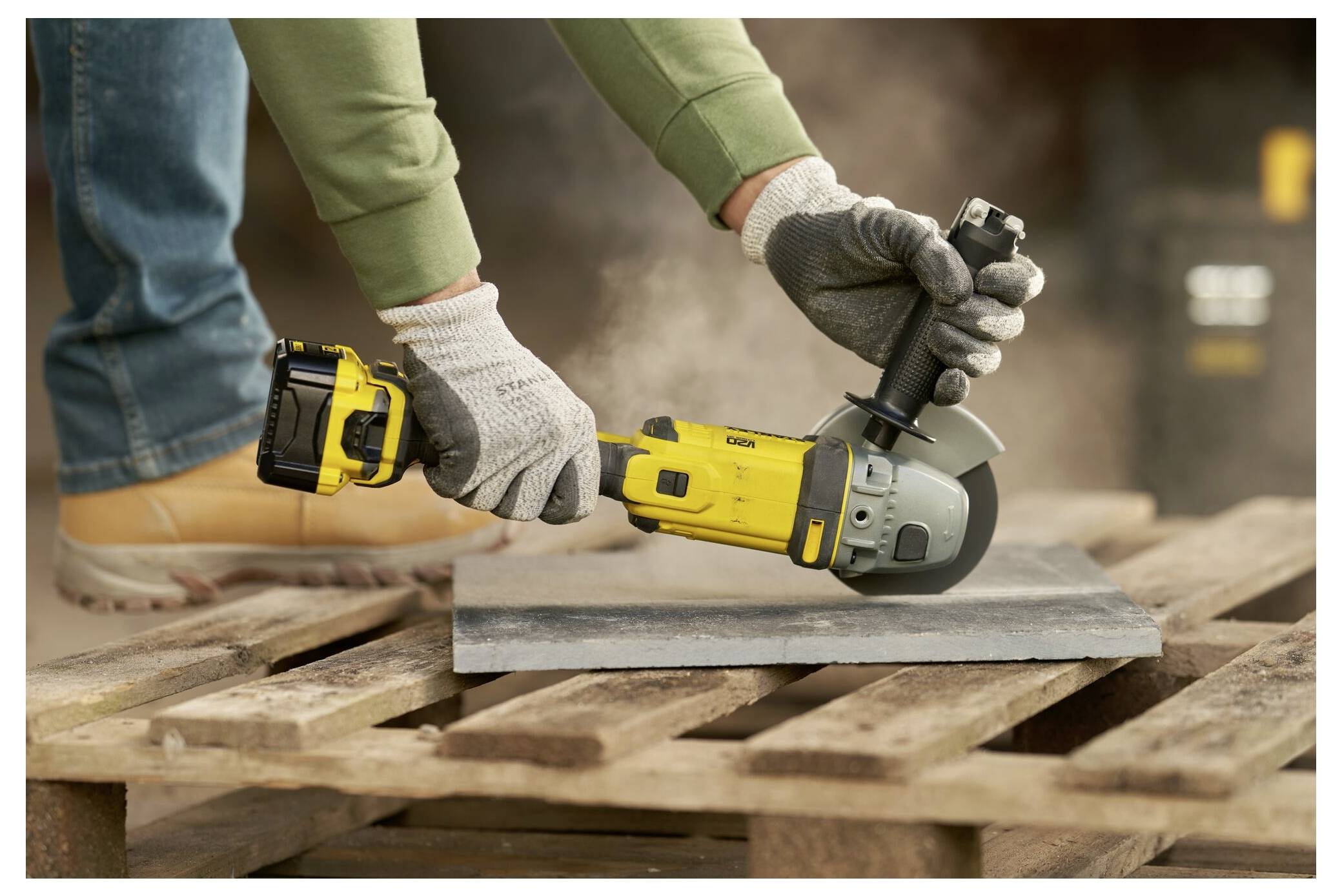 Eine Person verwendet einen gelben kabellosen Winkelschleiffer auf einem Metallblech, das auf einer hölzernen Palette liegt. Trägt Handschuhe und persönliche Schutzausrüstung.