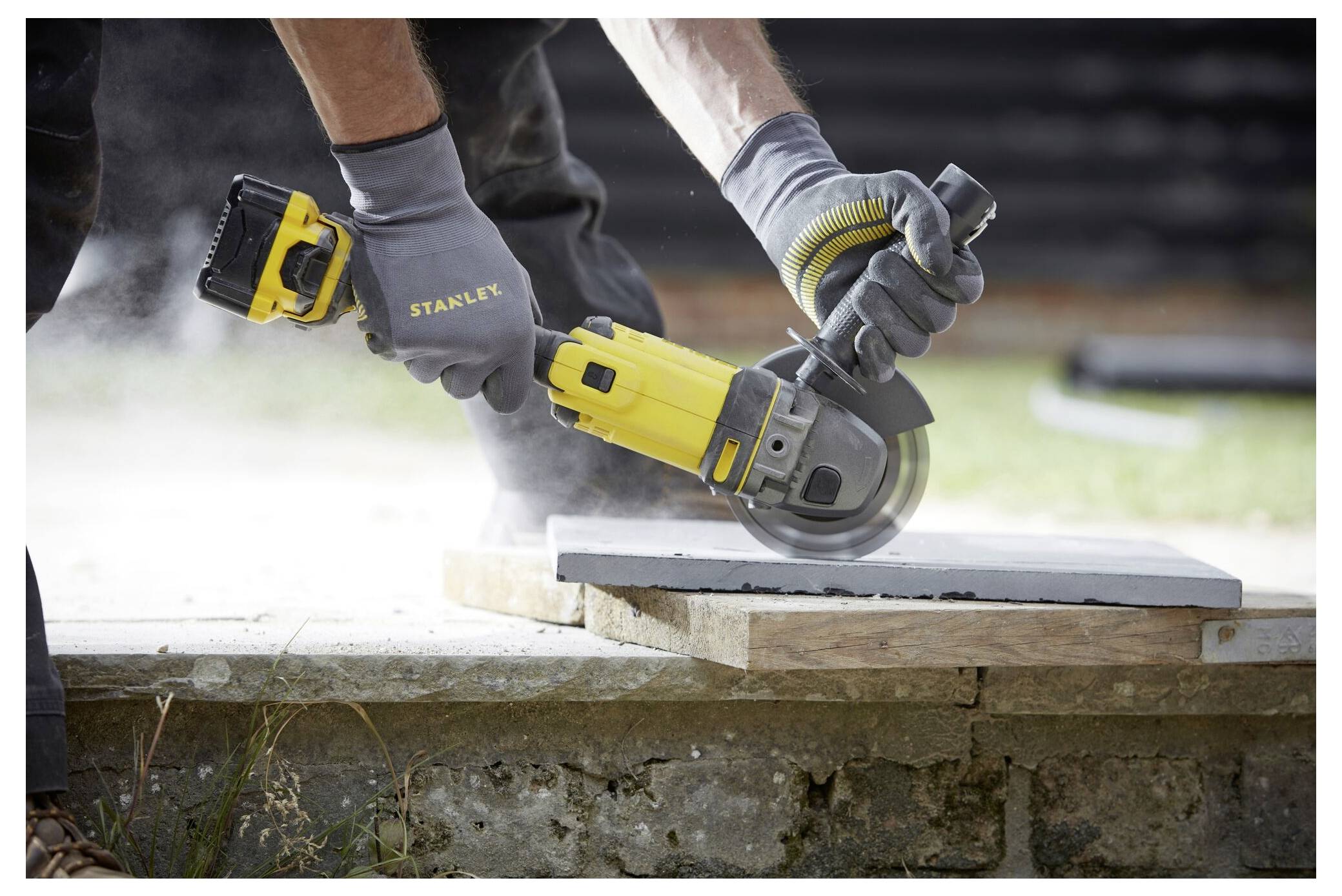 Eine Person mit Handschuhen schneidet mit einer batteriebetriebenen Kreissäge eine Betonplatte auf einer Baustelle, wobei Staub entsteht.