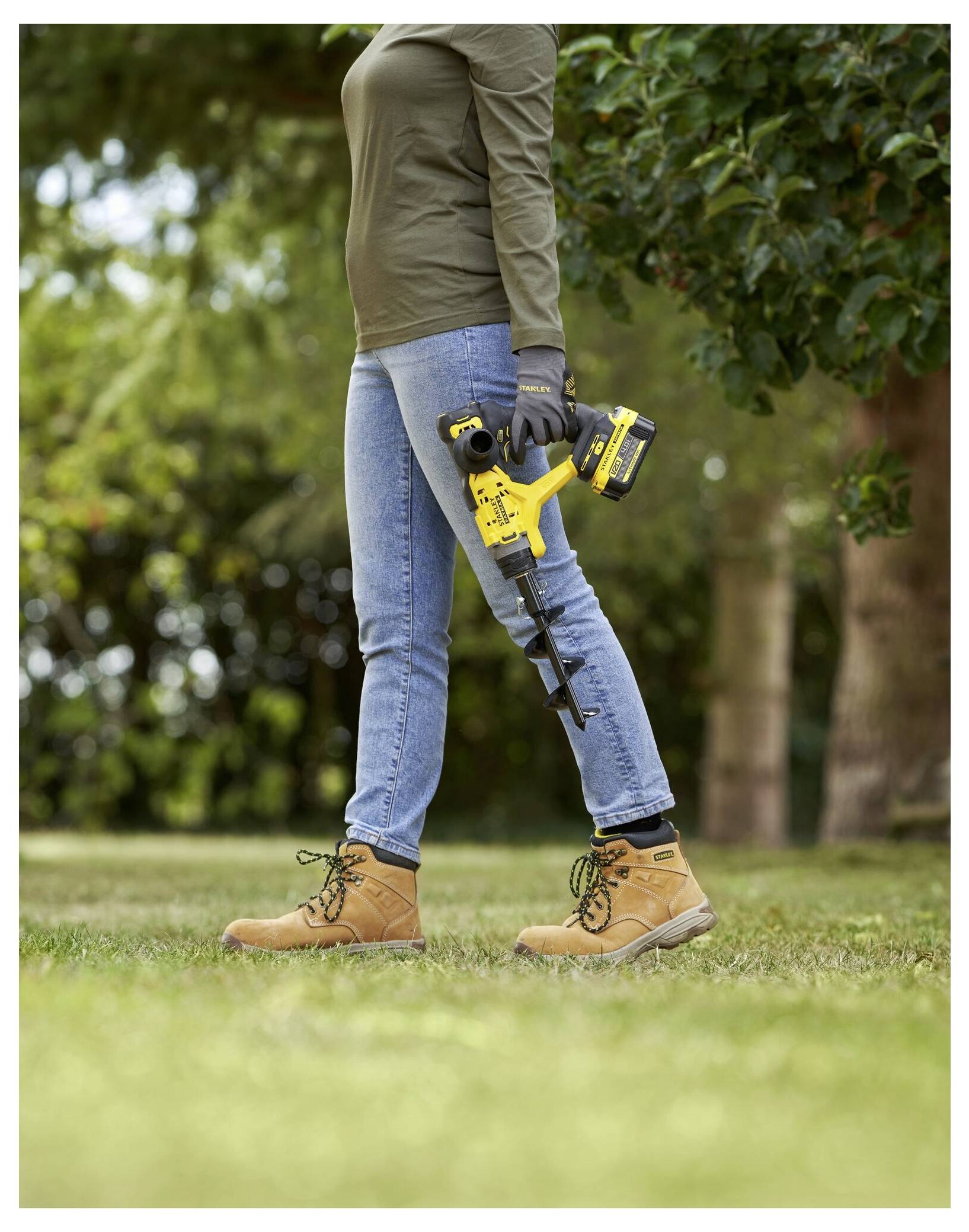 Person in casual attire holding a yellow cordless drill outdoors, surrounded by green trees and grass.Person in Freizeitkleidung, der einen gelben kabellosen Bohrer im Freien hält, umgeben von grünen Bäumen und Gras.