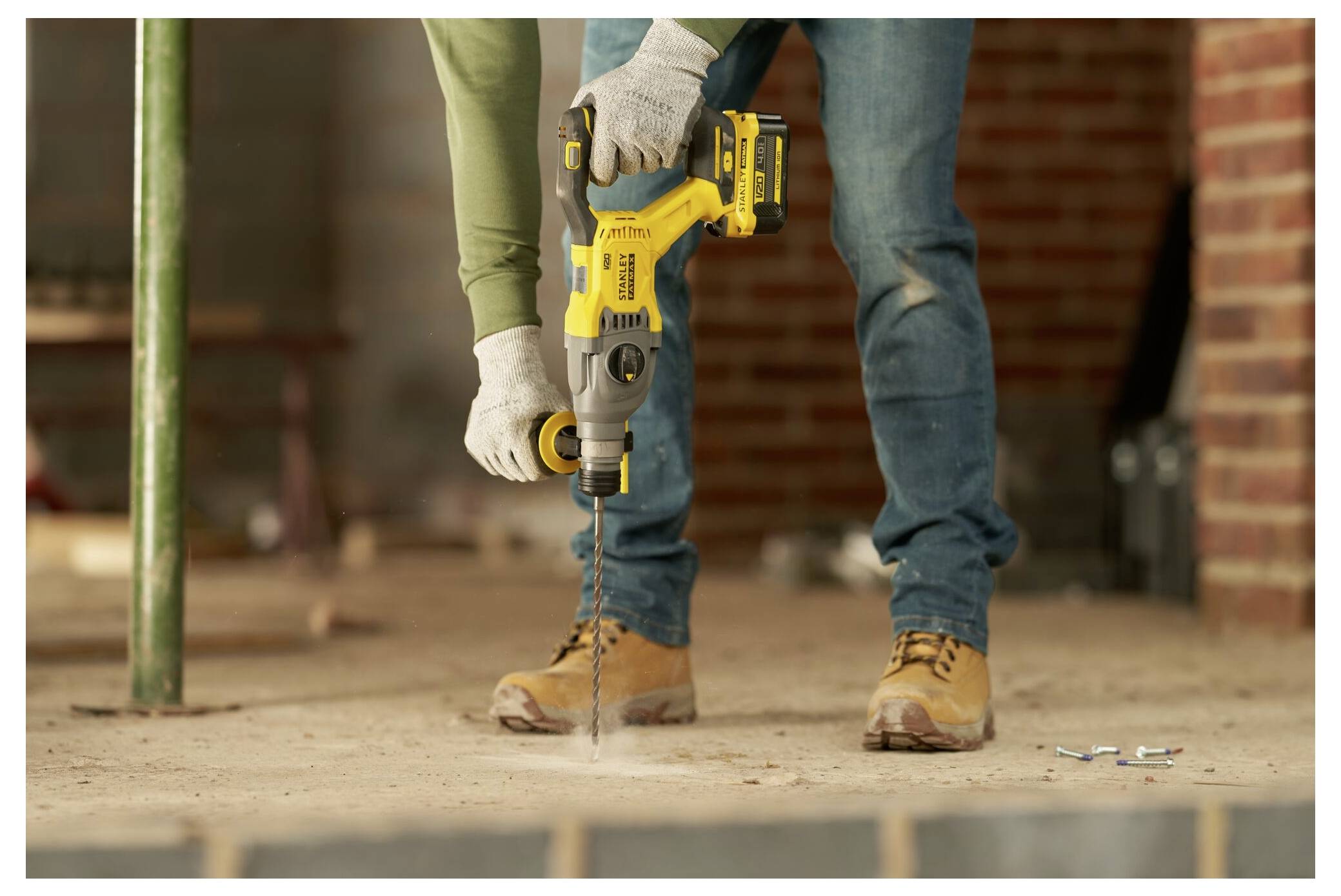 Eine Person in Arbeitsschuhen und Handschuhen benutzt eine gelbe Elektrobohrmaschine auf einem Betonboden in einem teilweise fertiggestellten Gebäude.