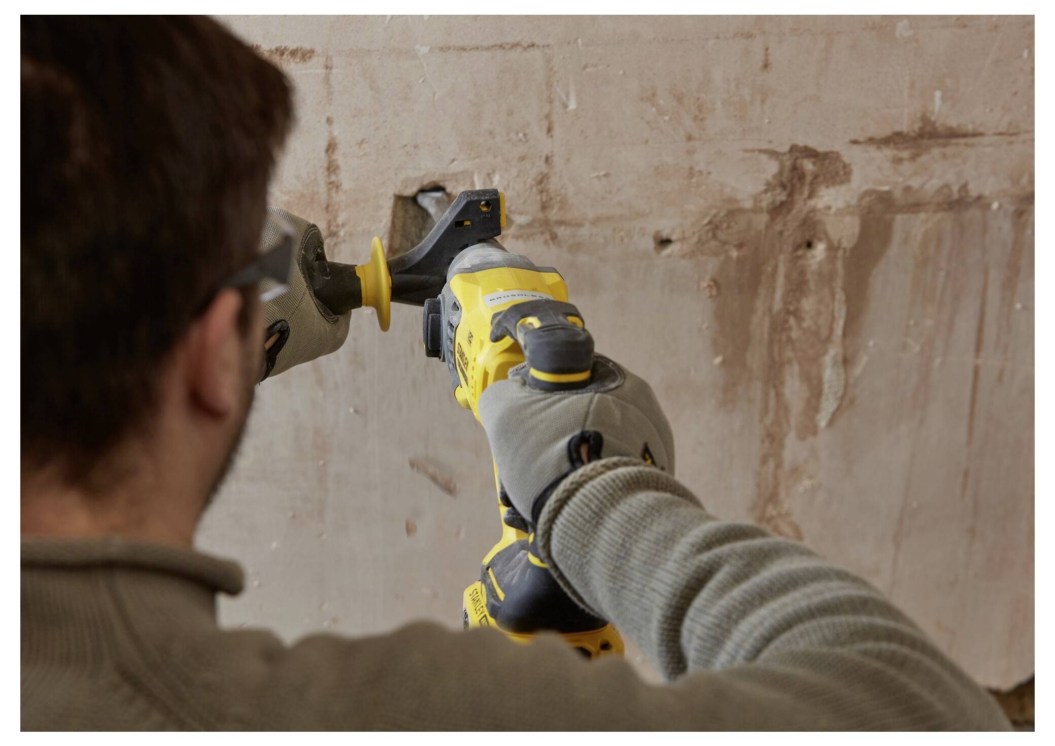 Eine Person, die Handschuhe trägt, bedient ein gelbes Elektrowerkzeug an einer Wand und scheint bei Bau- oder Abbrucharbeiten beschäftigt zu sein.