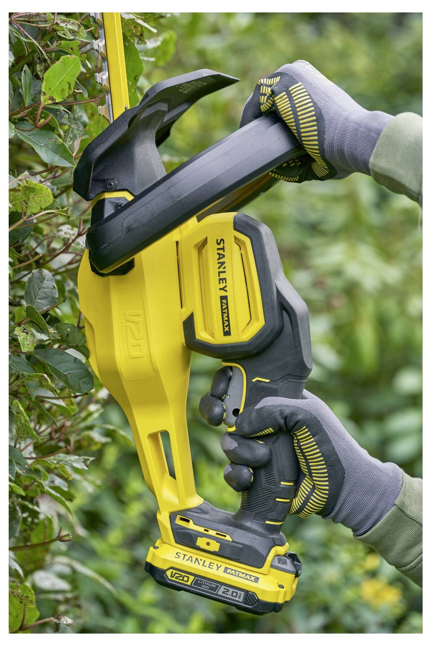 Eine Person, die eine gelb-schwarze elektrische Heckenschere der Marke Stanley trägt, schneidet einen Strauch, während sie Schutzhandschuhe trägt.