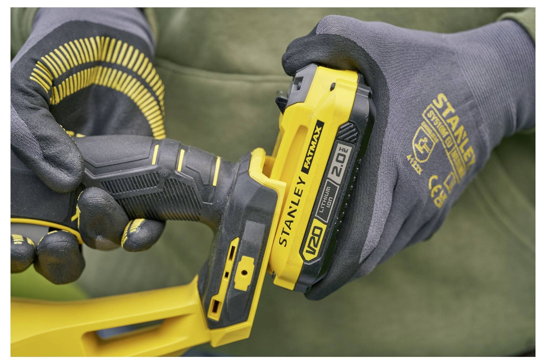 Eine Nahaufnahme von behandschuhten Händen, die eine Stanley V20 Lithium-Ionen-2,0-Ah-Batterie mit einem Elektrowerkzeug halten. Der Fokus liegt auf dem Batteriedetail.
