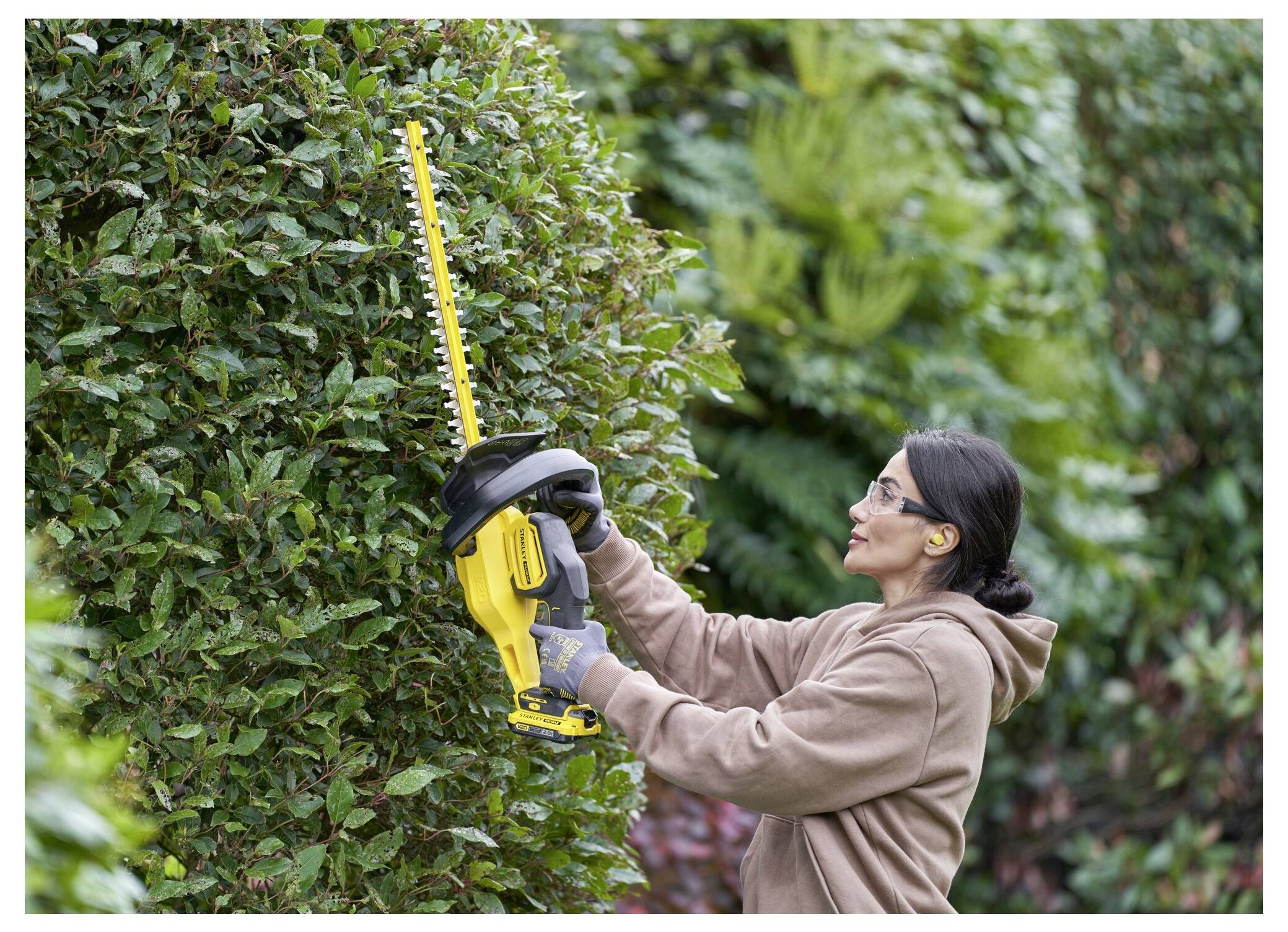 Eine Person, die Schutzbrille und Handschuhe trägt, verwendet eine elektrische Heckenschere, um einen großen Strauch in einem Garten zu formen.