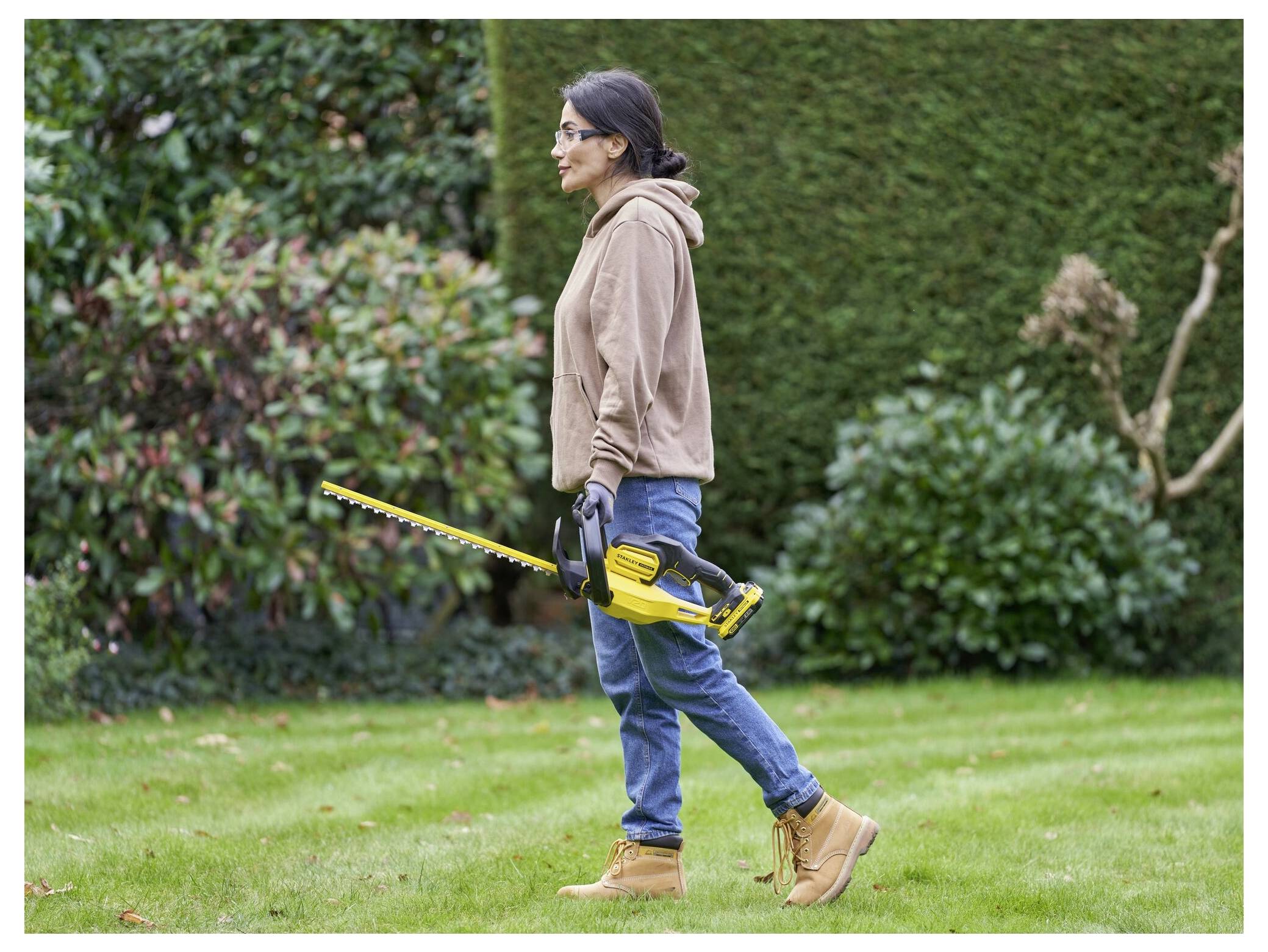 Eine Person, die eine Hecke in einem Garten mit einem gelben Elektrotrimmer schneidet, trägt einen braunen Hoodie, Jeans, Handschuhe und Stiefel.