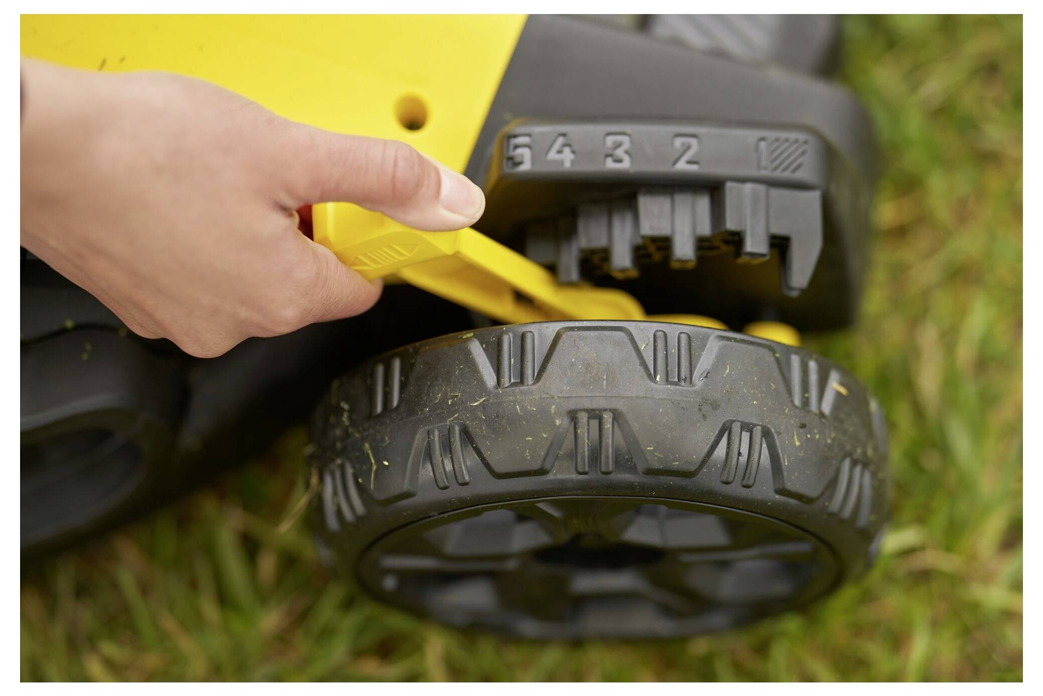 Eine Hand stellt die Höheneinstellung eines gelben Rasenmähers ein, wobei Zahlenmarkierungen über dem Rad auf Gras sichtbar sind.