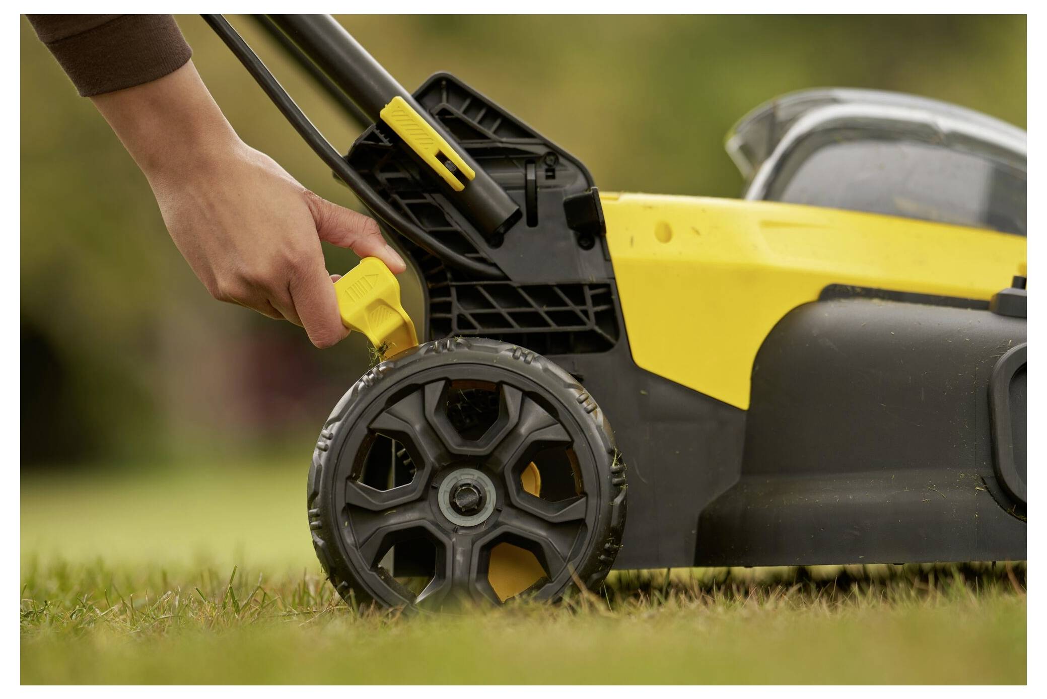 Eine Person stellt die Höhe eines gelb-schwarzen Rasenmäher auf Gras ein und konzentriert sich dabei auf den Rad- und Hebelmechanismus.