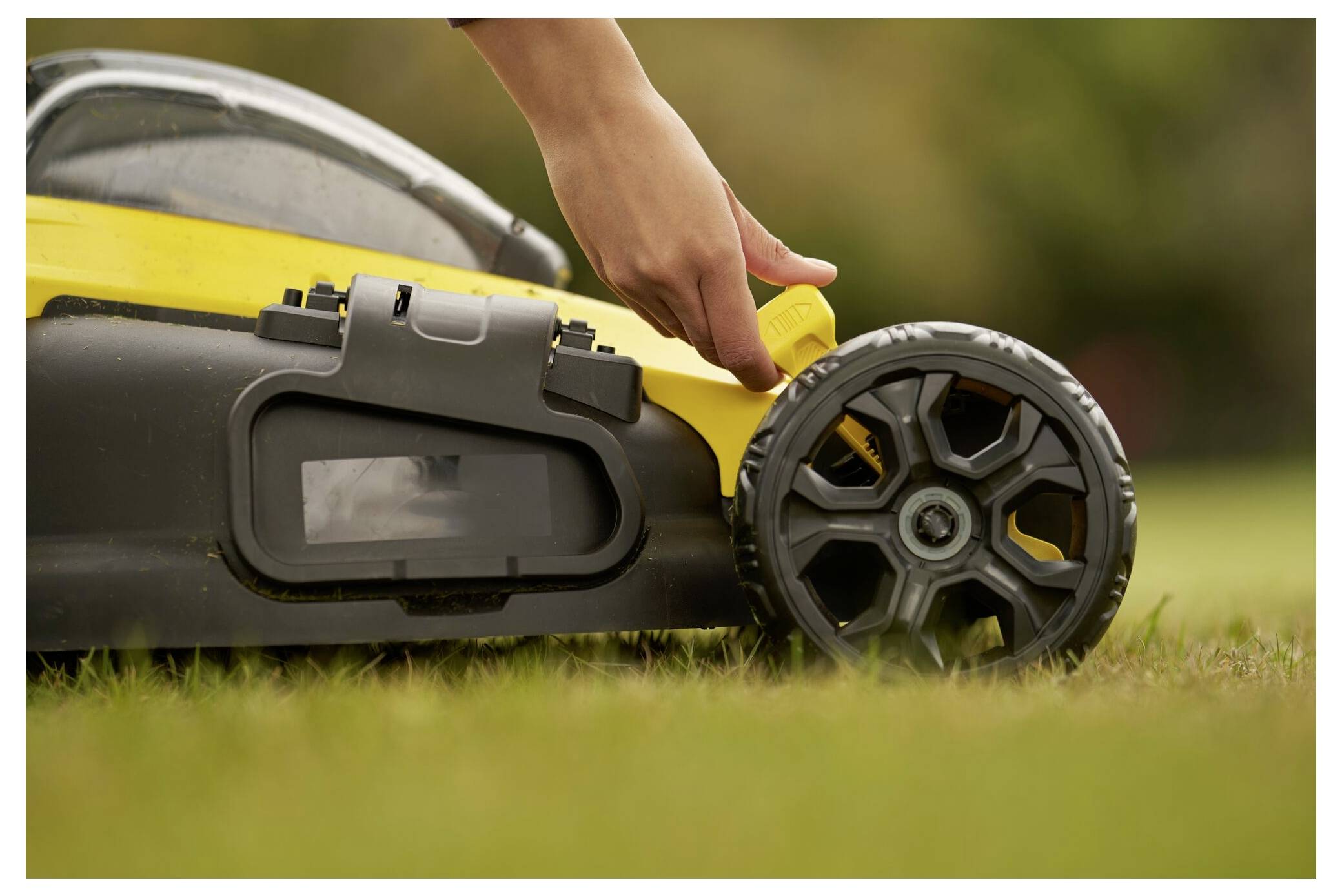 Eine Hand verstellt einen gelben Hebel an einem Rasenmäher und ändert dabei die Schnitthöheneinstellung der Räder. Der Hintergrund zeigt unscharfes grünes Gras.