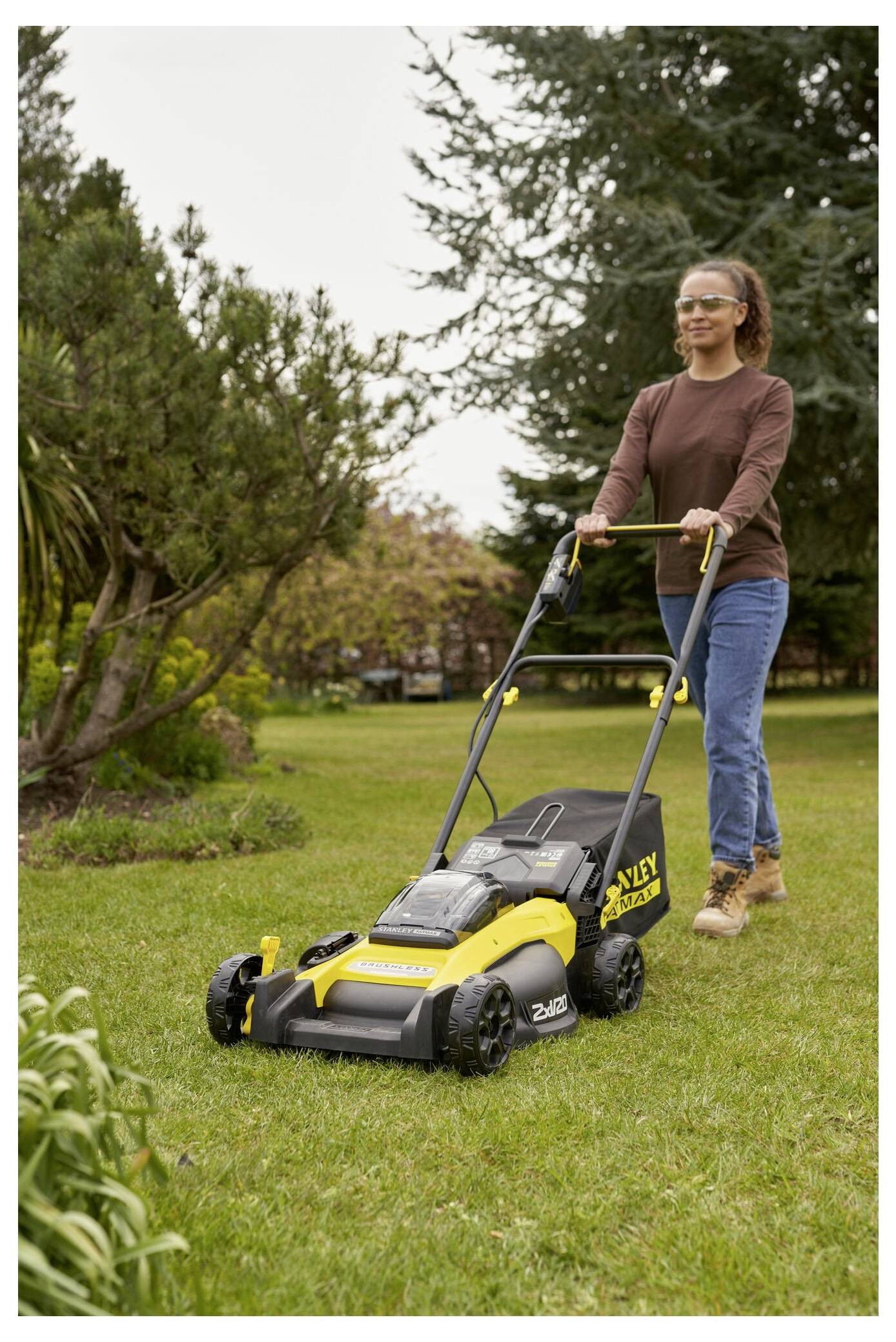 Eine Person mäht den Rasen mit einem gelb-schwarzen elektrischen Rasenmäher in einer Gartenumgebung. Bäume und Sträucher sind im Hintergrund sichtbar.