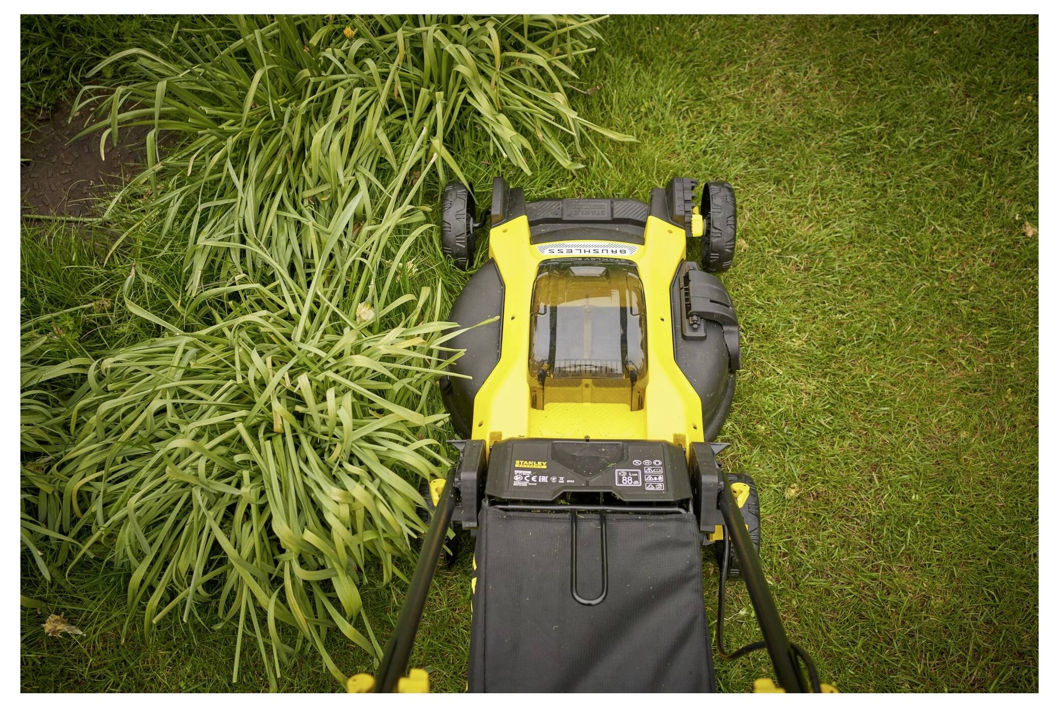 Ein gelber Rasenmäher wird von oben gezeigt, wie er durch ein dichtes Stück lange Gräser schneidet und links einen ordentlich gestutzten Weg hinterlässt.