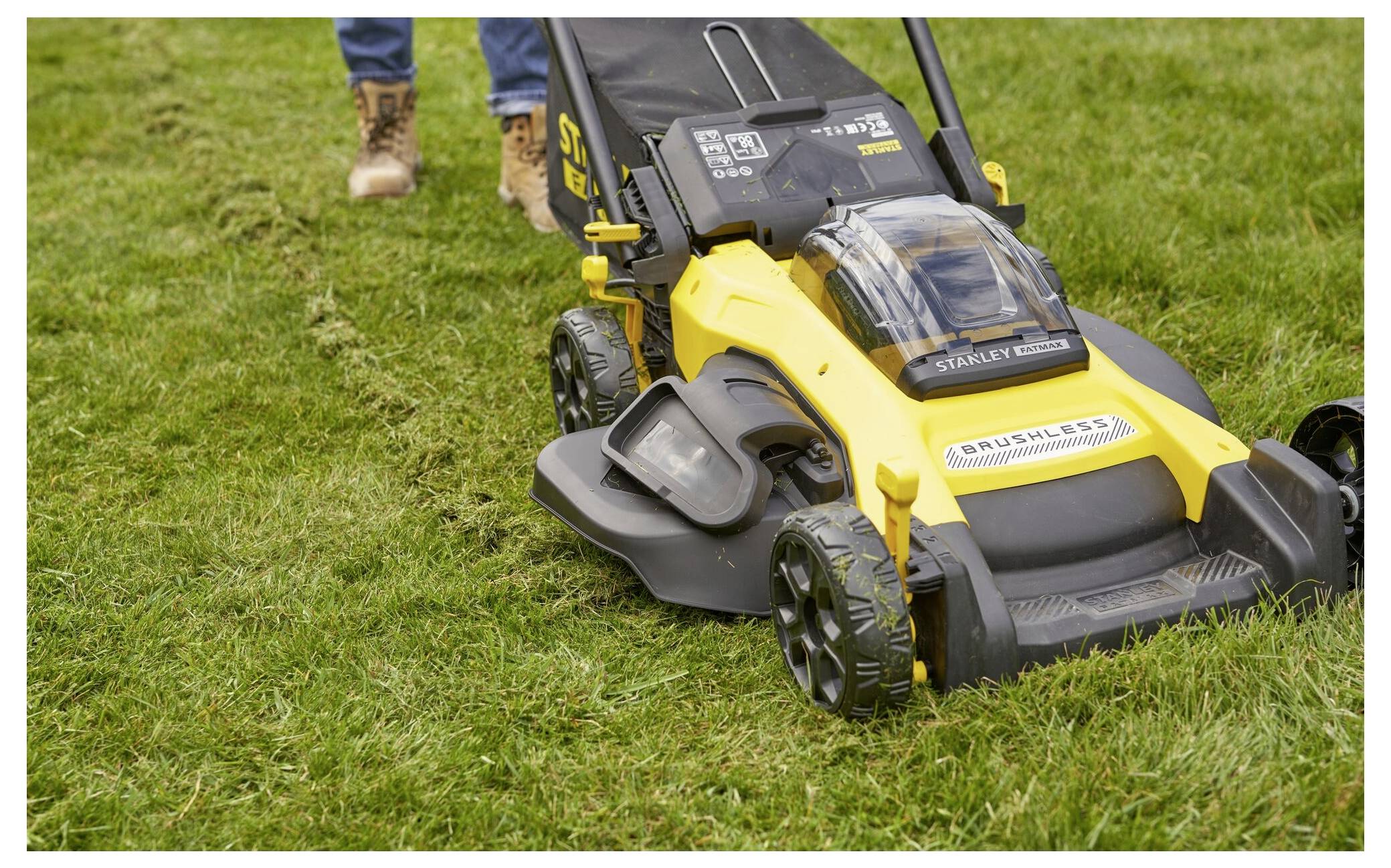 Eine Person benutzt einen gelb-schwarzen Rasenmäher auf einem Grasfläche, sichtbare Beine in Jeans und Stiefeln. Der Mäher wird mit Batterie betrieben.