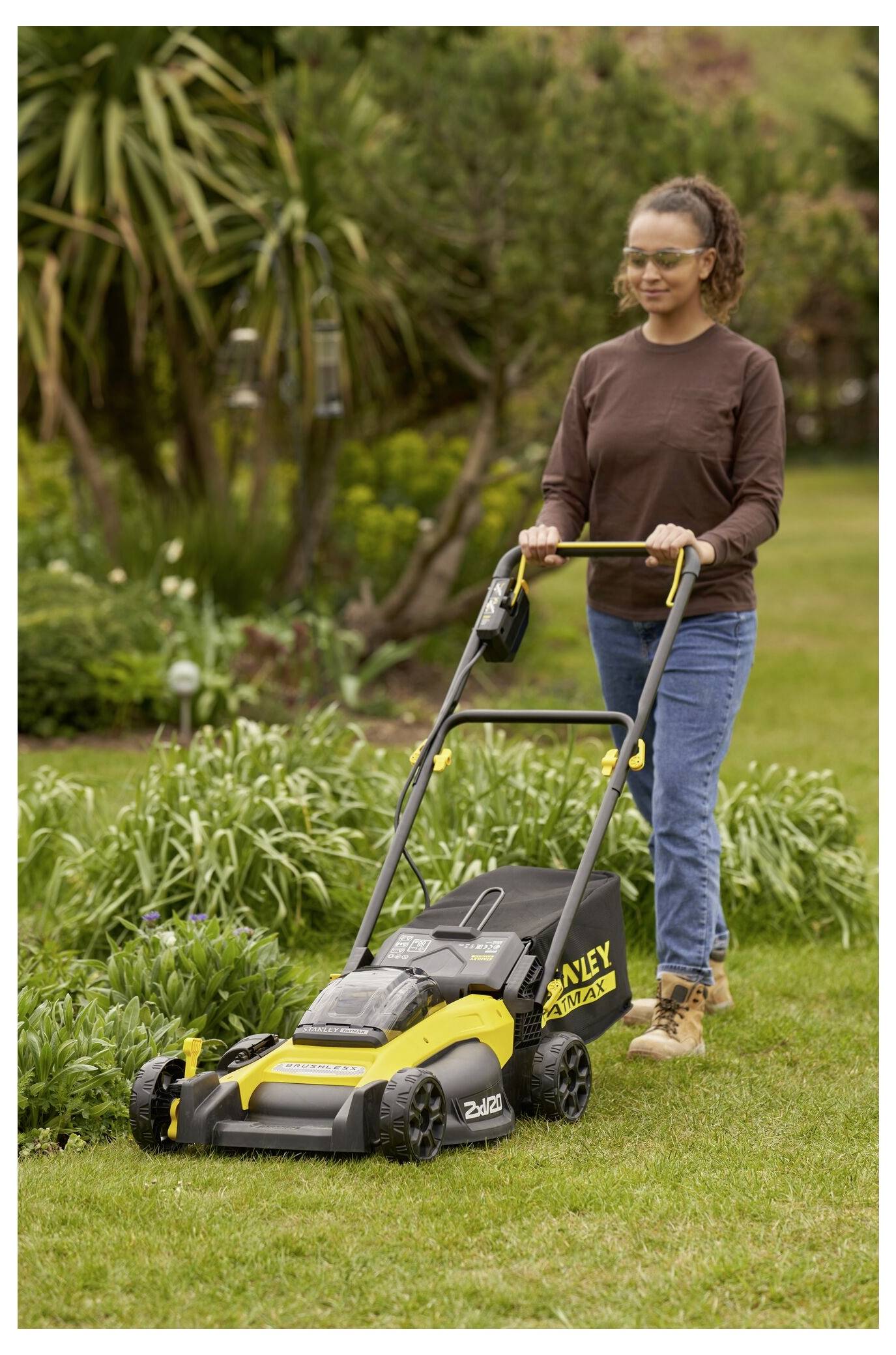 Eine Person, die ein braunes Langarm-Shirt und blaue Jeans trägt, mäht einen Rasen mit einem gelb-schwarzen Stanley-Akku-Rasenmäher.