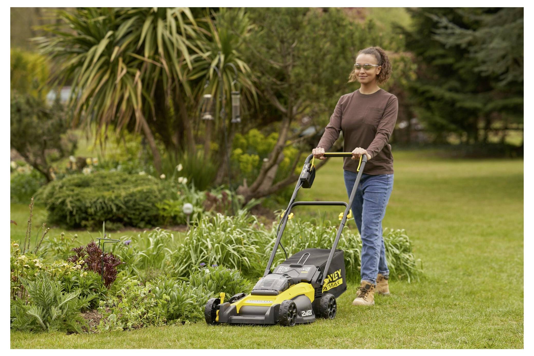 Eine Person mäht mit einem gelb-schwarzen Rasenmäher Gras in einem Garten, umgeben von Pflanzen und Bäumen unter einem klaren Himmel.