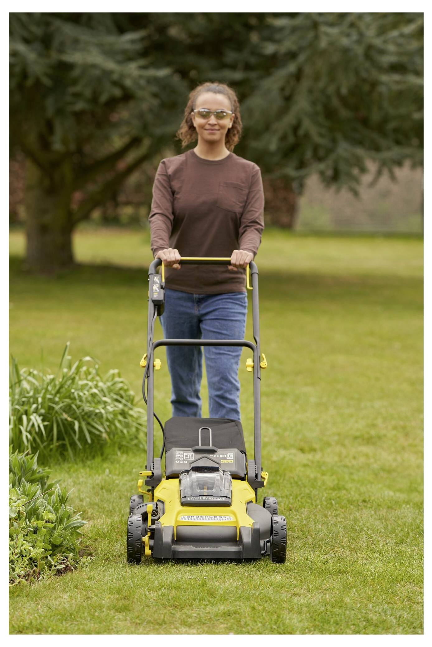 Eine Person, die mit einem gelb-schwarzen Rasenmäher den Rasen mäht, trägt Freizeitkleidung und Schutzbrille, mit Bäumen im Hintergrund.