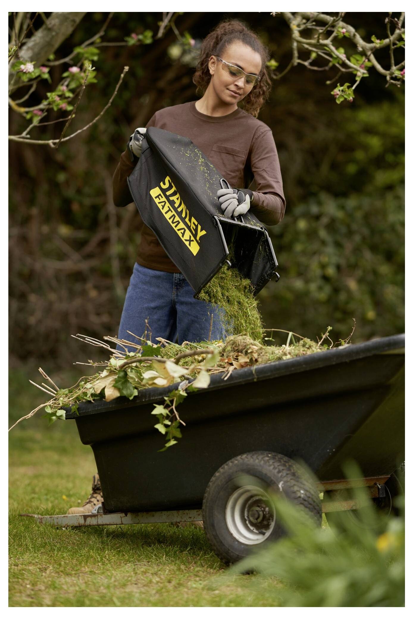 Eine Frau, die Handschuhe trägt, schüttet Grasschnitt und Blattwerk aus einem schwarzen „Stanley Fatmax