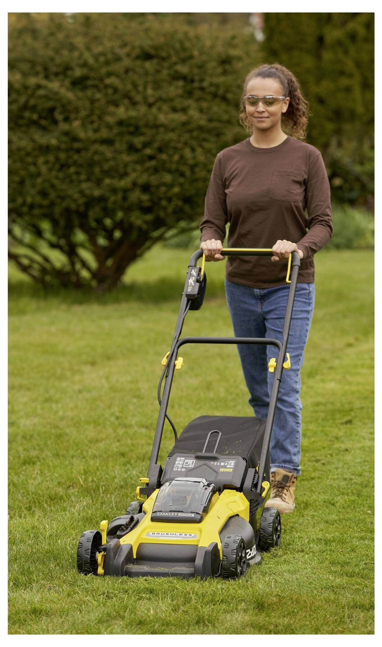 Eine Person mit Sonnenbrille schiebt einen gelben Rasenmäher auf einem Rasen. Im Hintergrund sind Büsche und Bäume sichtbar.