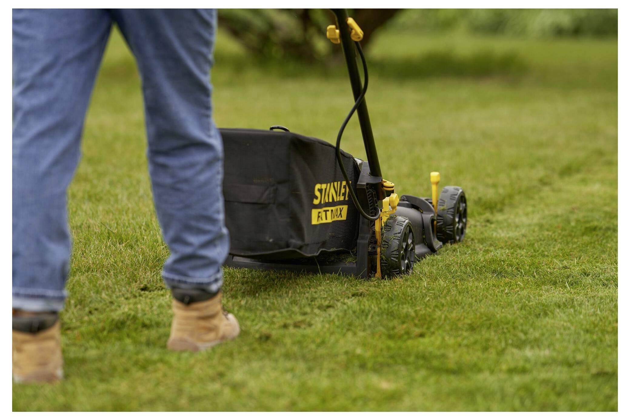 Eine Person mäht einen Rasen mit einem Stanley-Rasenmäher. Die Person trägt Jeans und Stiefel und konzentriert sich darauf, das Gras gleichmäßig zu schneiden.