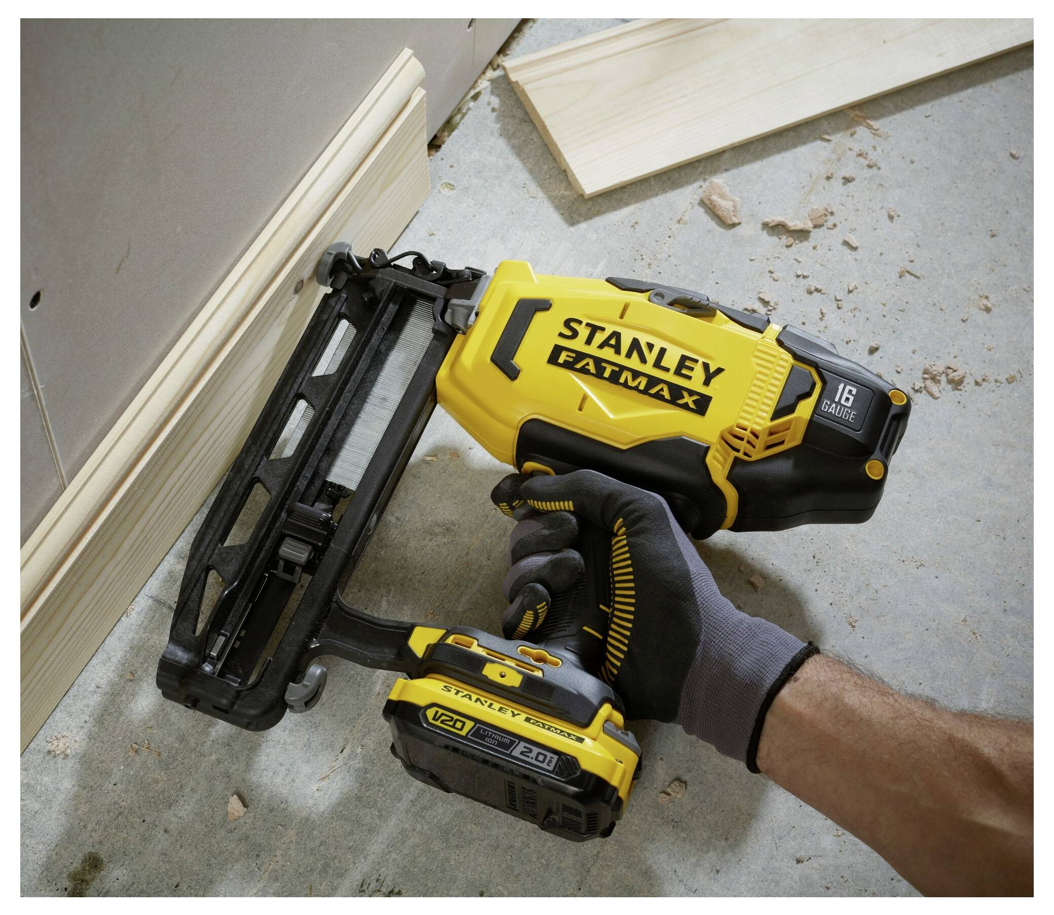 Eine Person mit Handschuhen benutzt eine Stanley Fatmax Nagelschussmaschine, um ein Holzbrett an einer Wand zu befestigen. Werkzeuge und Holzspäne sind sichtbar.